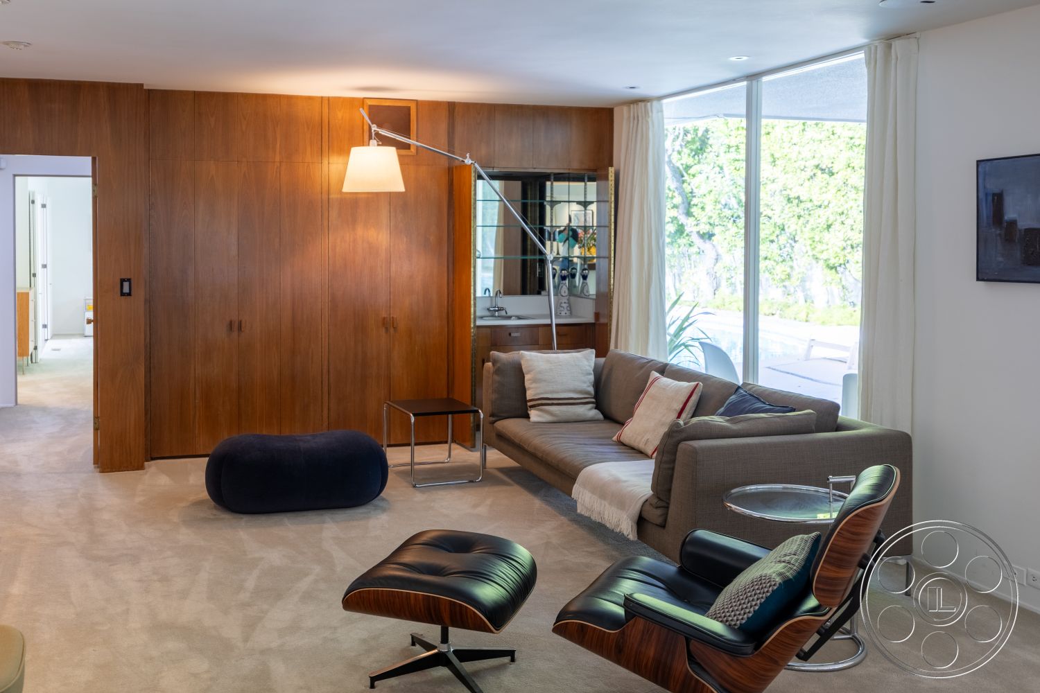 MCM 27 - integrated lighting fixtures, contemporary design aesthetics, open floor plan living area, glass floor-to-ceiling windows, minimalist coffee table design, elegant curtain drapery, mid-century modern armchair, modern wall art decor, architectural details with clean lines, walnut wood paneling interior, contemporary sofa with neutral upholstery, soft beige carpet flooring, suburban setting with garden view, comfort-focused seating arrangement, casual yet sophisticated living space