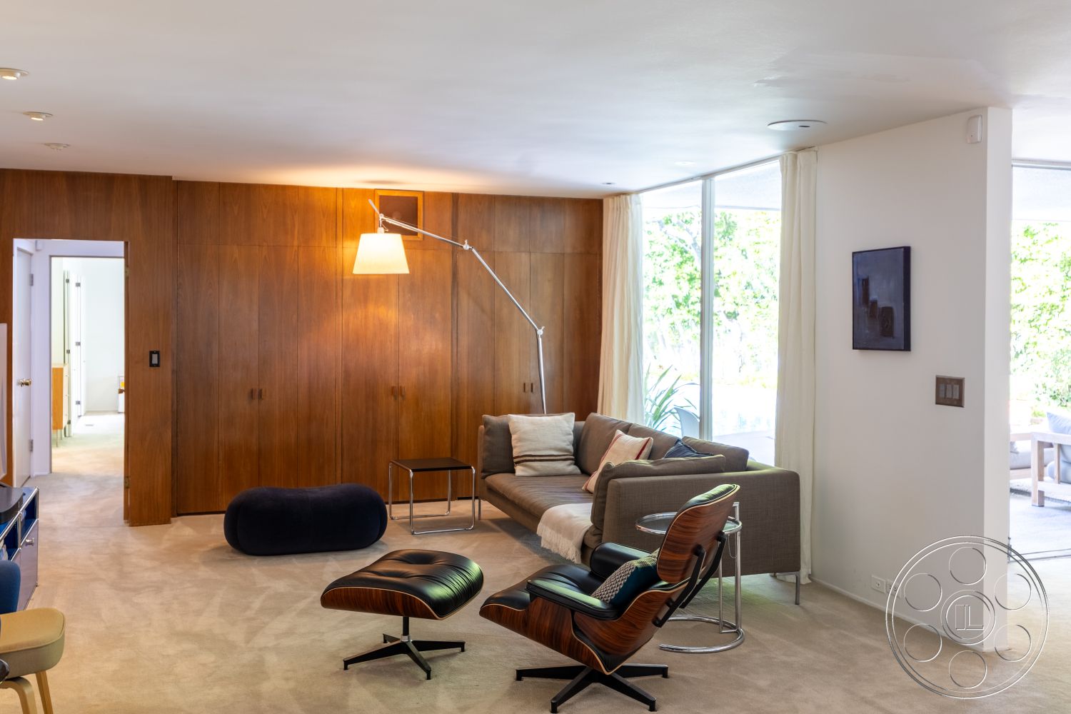 MCM 27 - neutral color palette, open floor plan, glass coffee table, outdoor access, floor-to-ceiling windows, contemporary design elements, mid-century modern style, white sheer curtains, decorative floor lamp, modern sectional sofa, exposed wood wall panels, minimalist interior, plush carpet flooring, soft textured cushions, sitting area with natural light