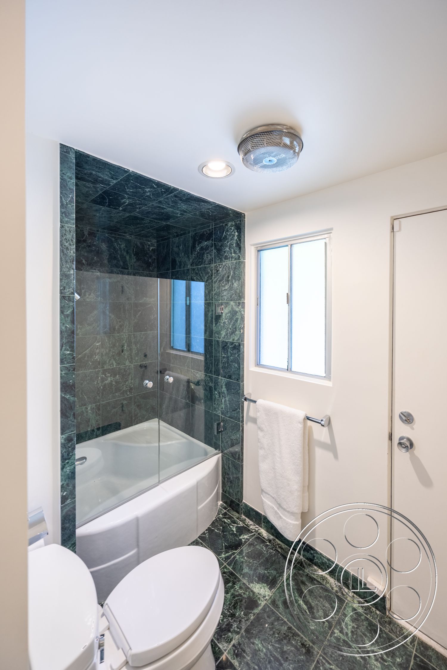 MCM 27 - glass shower enclosure, painted white walls, flush mount ceiling light, interior bathroom, toilet with soft-close lid, chrome bathroom fixtures, green marble bathroom flooring, green marble shower walls, white porcelain bathtub, white towel rack, window with white frame, modern vanity door