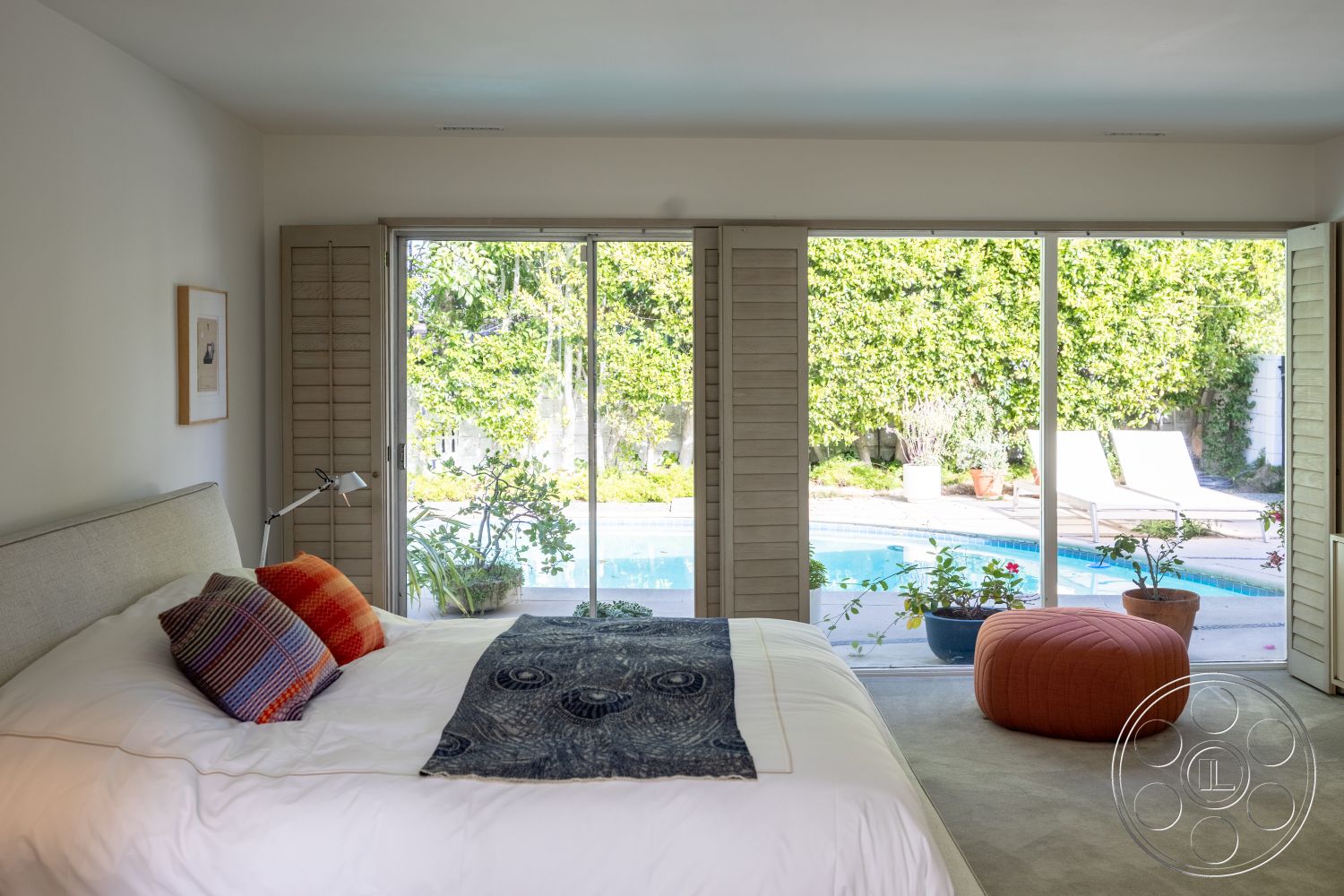 MCM 27 - contemporary design elements, decorative throw pillows, outdoor pool view, carpet bedroom flooring, textured white walls, natural light from sliding doors, interior bedroom with large glass sliding doors, light wood shutters, modern ottoman seating, plant accents in window space, minimalistic furniture design, greenery in backyard setting, sunlit room ambiance