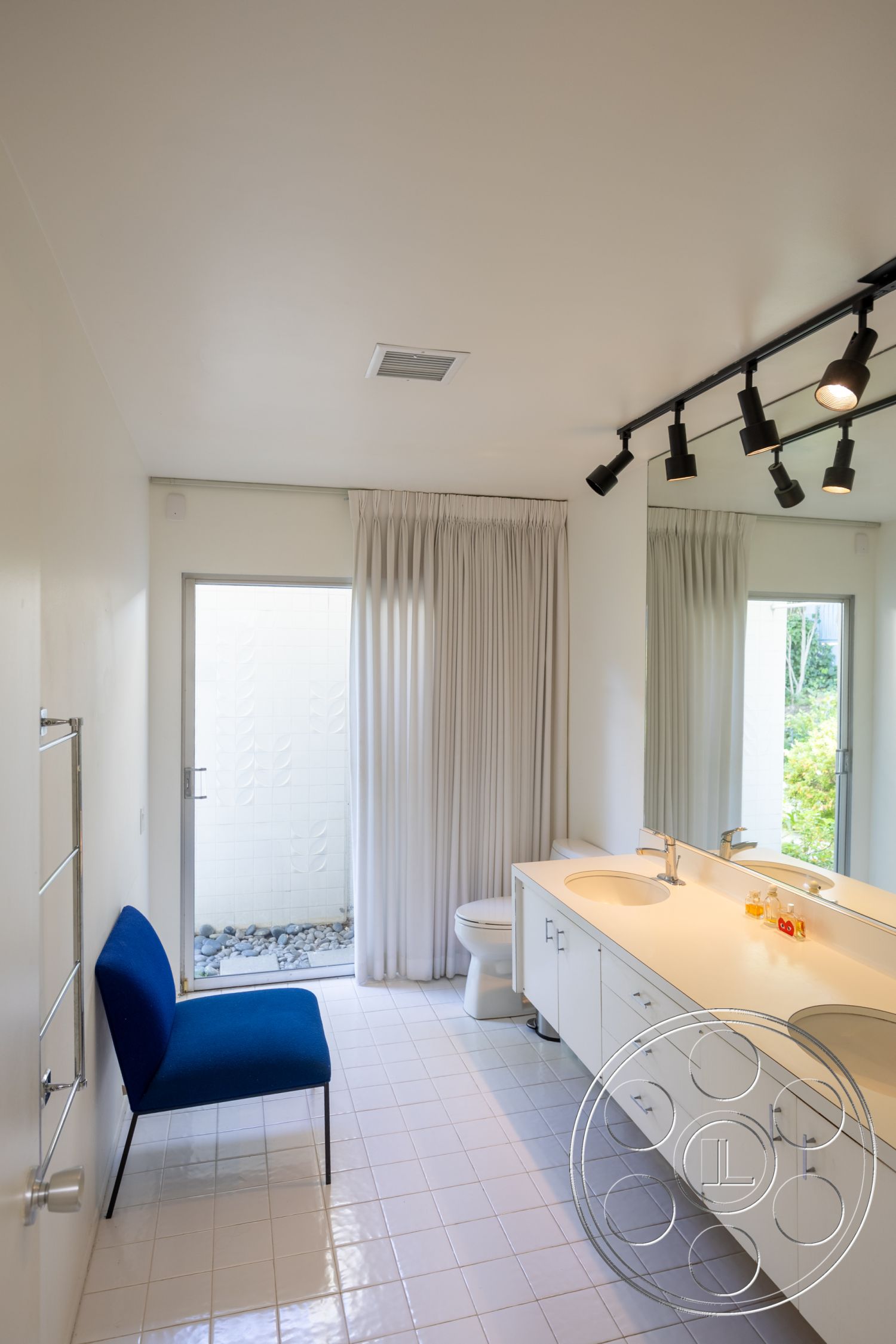 MCM 27 - glass shower enclosure, blue accent chair, white painted walls, ceramic tile bathroom walls, framed mirror above vanity, interior bathroom with tile flooring, modern fixtures and cabinetry, double vanity sink with quartz countertop, floor-to-ceiling window with sheer curtains, pebble stone landscaping visible through window, track lighting for contemporary ambiance