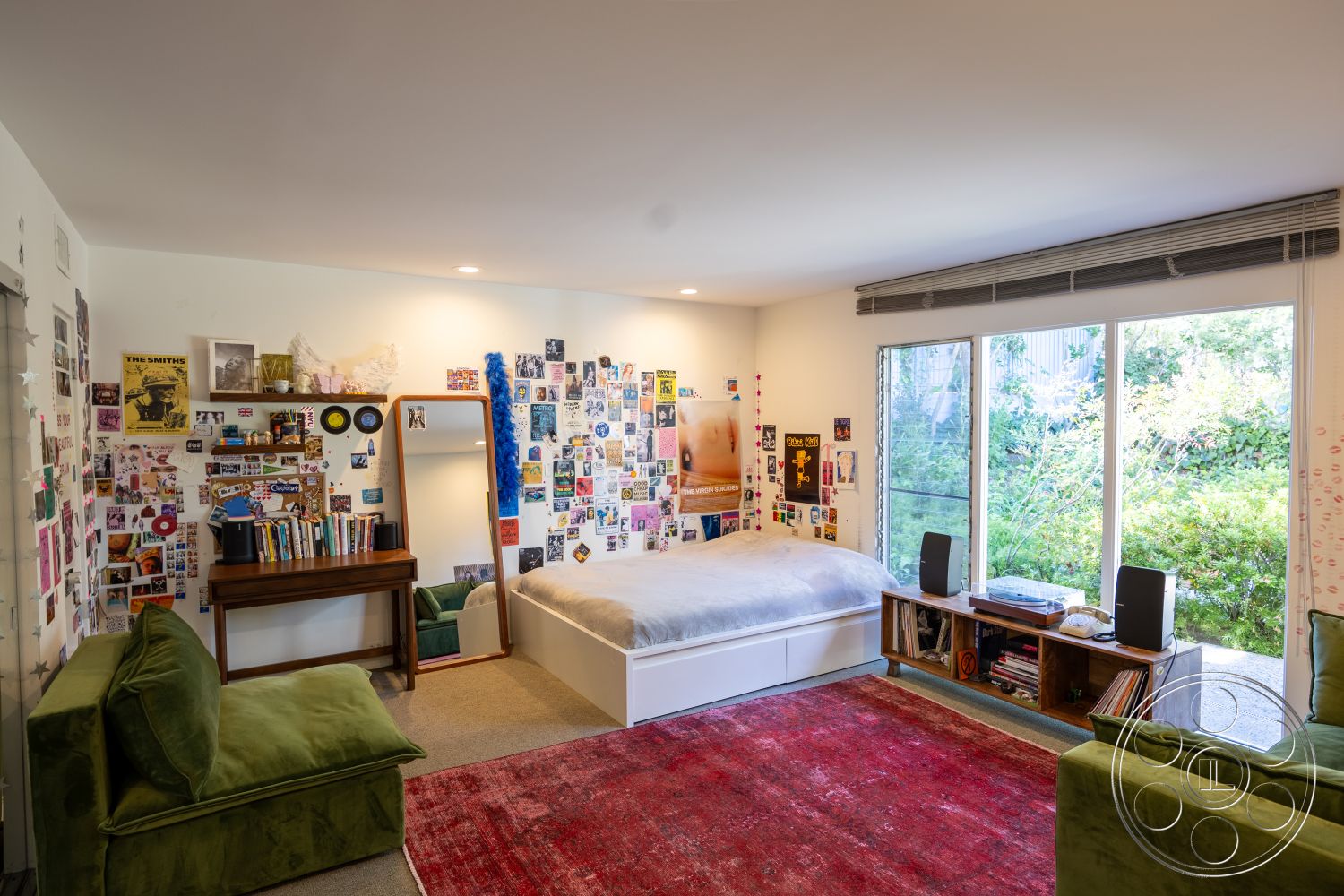 MCM 27 - open floor plan, decorative mirror, minimalist design elements, interior bedroom, textured carpet flooring, white painted walls, large glass sliding windows, plush green sofa, rectangular bed frame, wooden vanity table, view of outdoor garden, colorful wall art collage, natural light decor, cozy rug flooring, modern interior styling