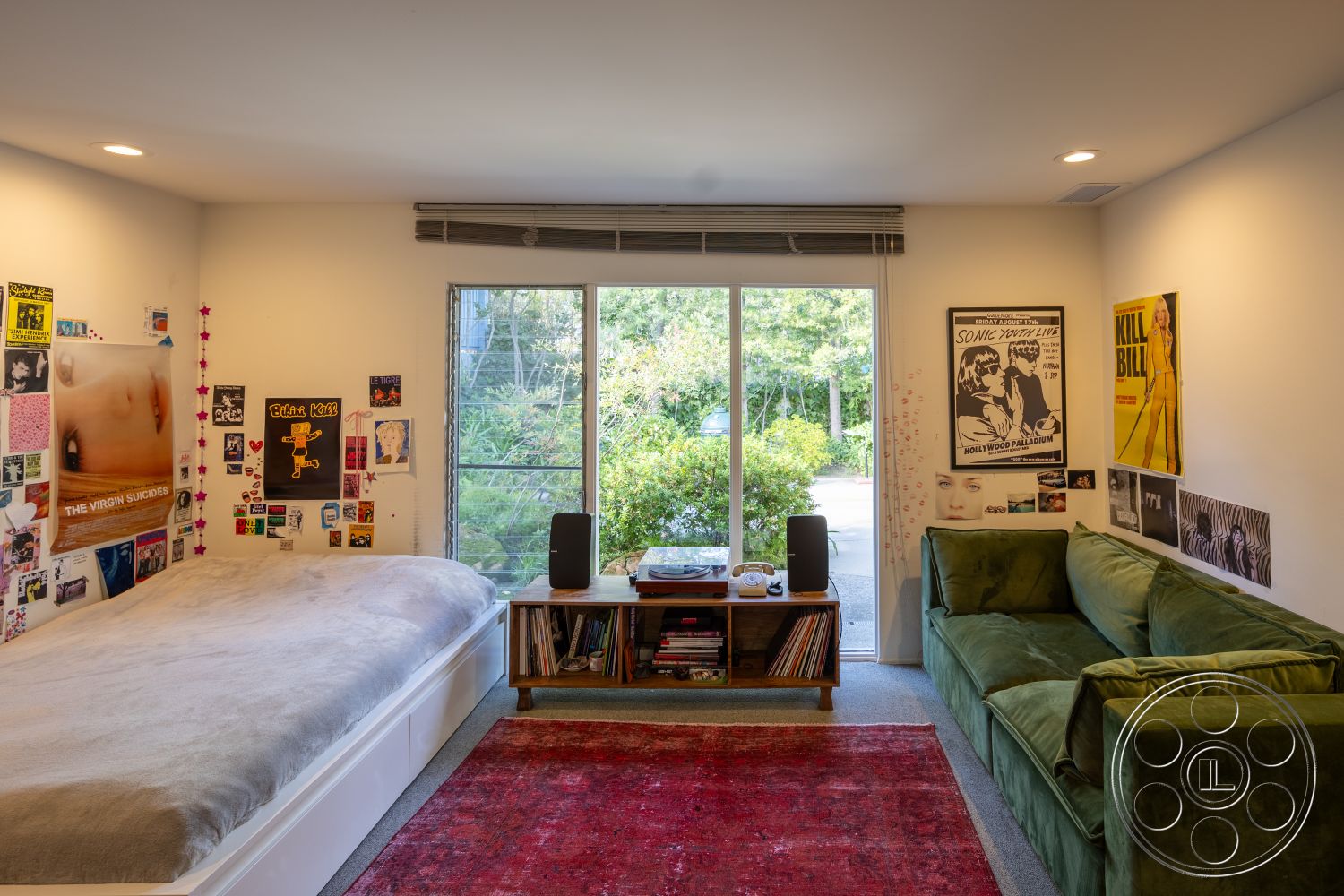 MCM 27 - neutral color palette, textured area rug, minimalist design elements, wooden furniture accents, soft bedding materials, large glass sliding windows, interior bedroom with carpet flooring, contemporary style room, white painted walls with wall art, plush green velvet sofa, decorative posters and photographs, natural light from exterior garden view, record storage unit with vinyl records, stylish bedside decor, understated modern aesthetic