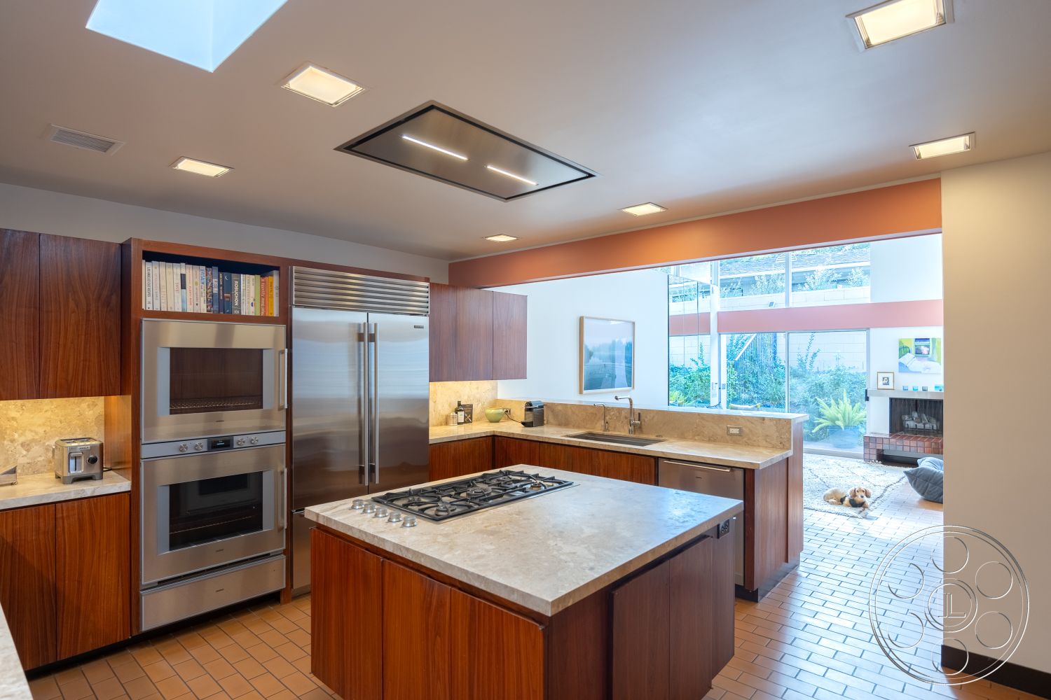 MCM 27 - contemporary kitchen design, natural light through large windows, minimalist aesthetic with clean lines, glass sliding doors leading to outdoor space, stainless steel appliances in modern kitchen, exterior kitchen with wood cabinetry, interior concrete countertops, interior tile flooring in warm tones, open layout chef's kitchen, exposed wood beams in ceiling, built-in oven and microwave in cabinetry, wood veneer cabinet fronts, kitchen island with gas cooktop, skylight above kitchen island, patio view from kitchen, outdoor garden setting visible