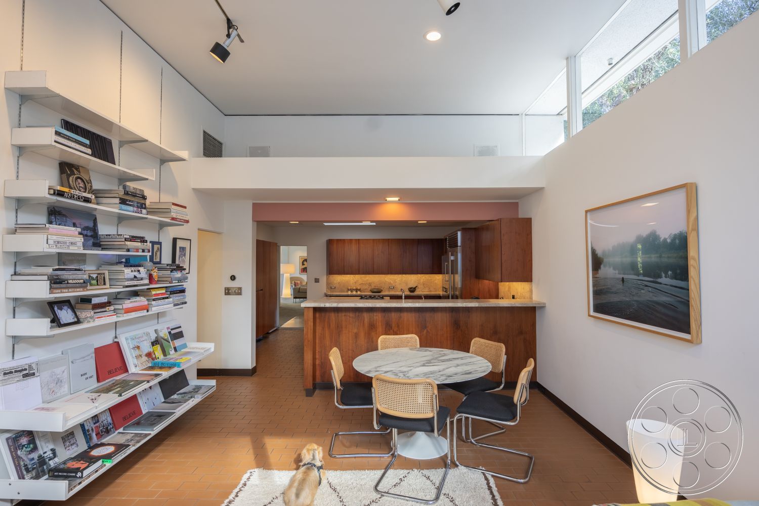 MCM 27 - contemporary decor, textured area rug, minimalist design elements, artwork on walls, large windows for natural light, interior space, round dining table, bright and airy atmosphere, cozy reading nook, terracotta floor tiles, marble kitchen countertop, white painted walls, open floor plan living area, high ceilings with recessed lighting, modern kitchen with wood cabinets, exposed shelving for books, metal and wood dining chairs, functional kitchen with modern appliances, seamless transition from kitchen to dining area, living room with dog, simple furniture arrangement