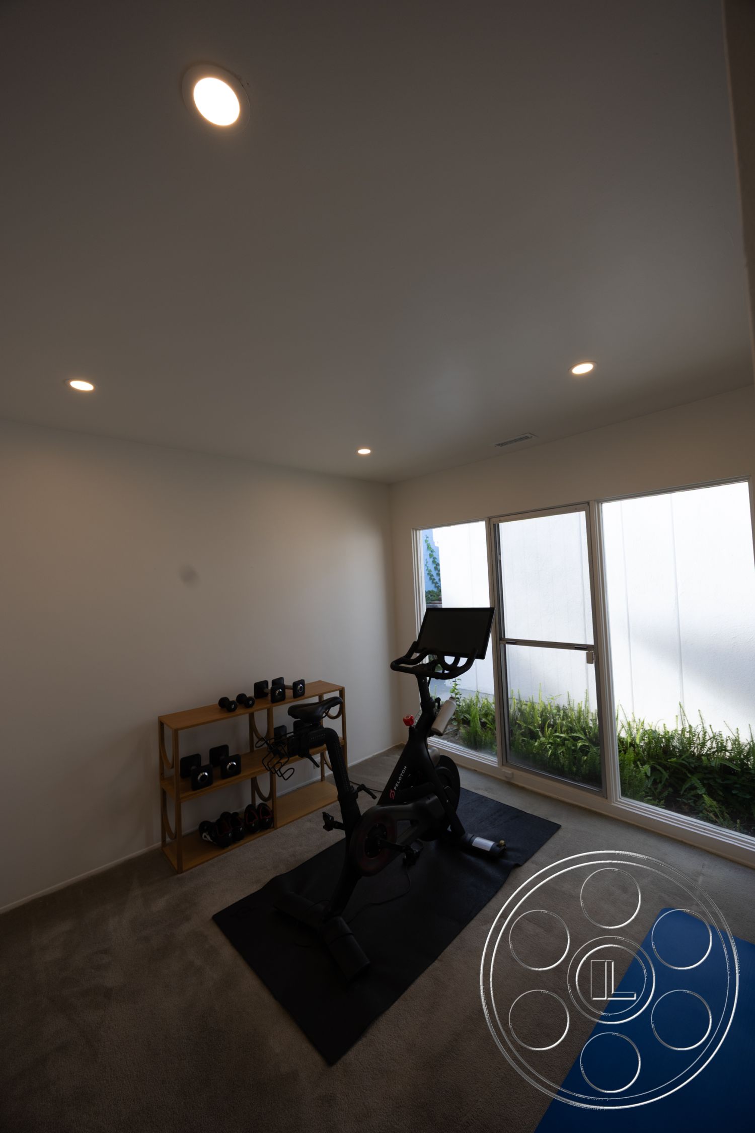 MCM 27 - floor-to-ceiling windows, recessed lighting fixtures, minimalist interior design, neutral painted walls, carpet flooring in fitness room, wall-mounted exercise bike, wooden storage shelf for weights, exercise mat on carpet, indoor workout space with natural light, functional home gym design