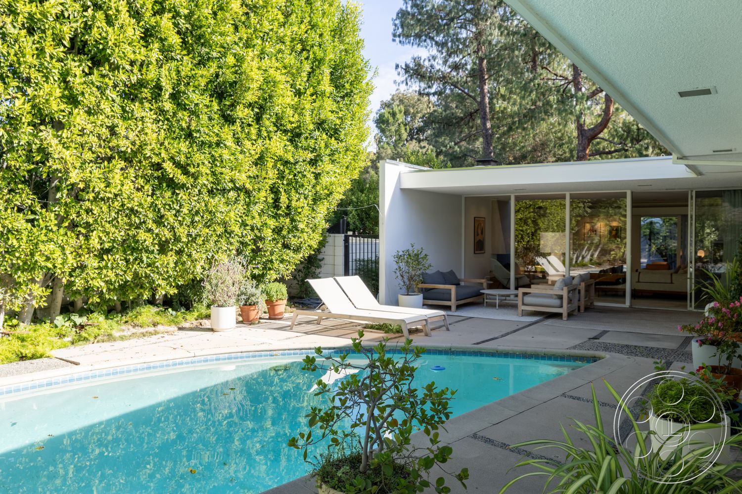 MCM 27 - open floor plan, sliding glass doors, floor-to-ceiling windows, rectangular pool, outdoor patio space, lush green landscaping, mid-century modern architectural style, suburban backyard setting, concrete patio floor, natural light-filled spaces, white stucco exterior walls, interior living area, surrounding pine trees, wooden sun loungers, exposed aggregate pool coping, minimalistic outdoor lounge area, container plants