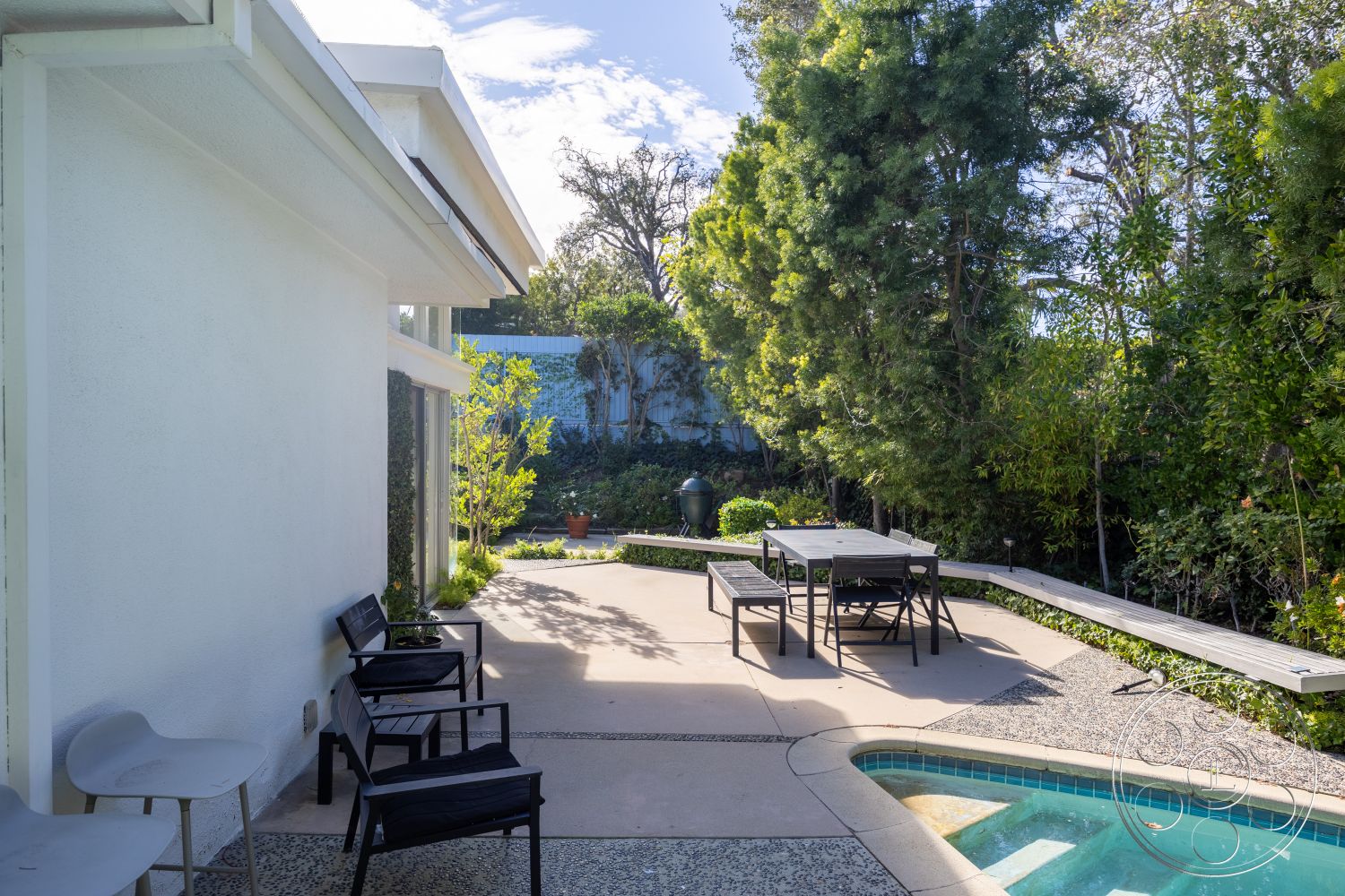 MCM 27 - white stucco walls, contemporary outdoor furniture, sunny outdoor space, exterior concrete patio floor, stone pathway edging, urban garden patio, in-ground rectangular swimming pool, wooden dining table with black metal chairs, landscaped garden setting with shrubs and greenery, comfortable outdoor seating area, modern design patio layout, tree-lined backyard environment, flat roof with overhang, geometric shaped pool, paved pool deck with decorative pattern, outdoor dining area for entertaining, open space for relaxation