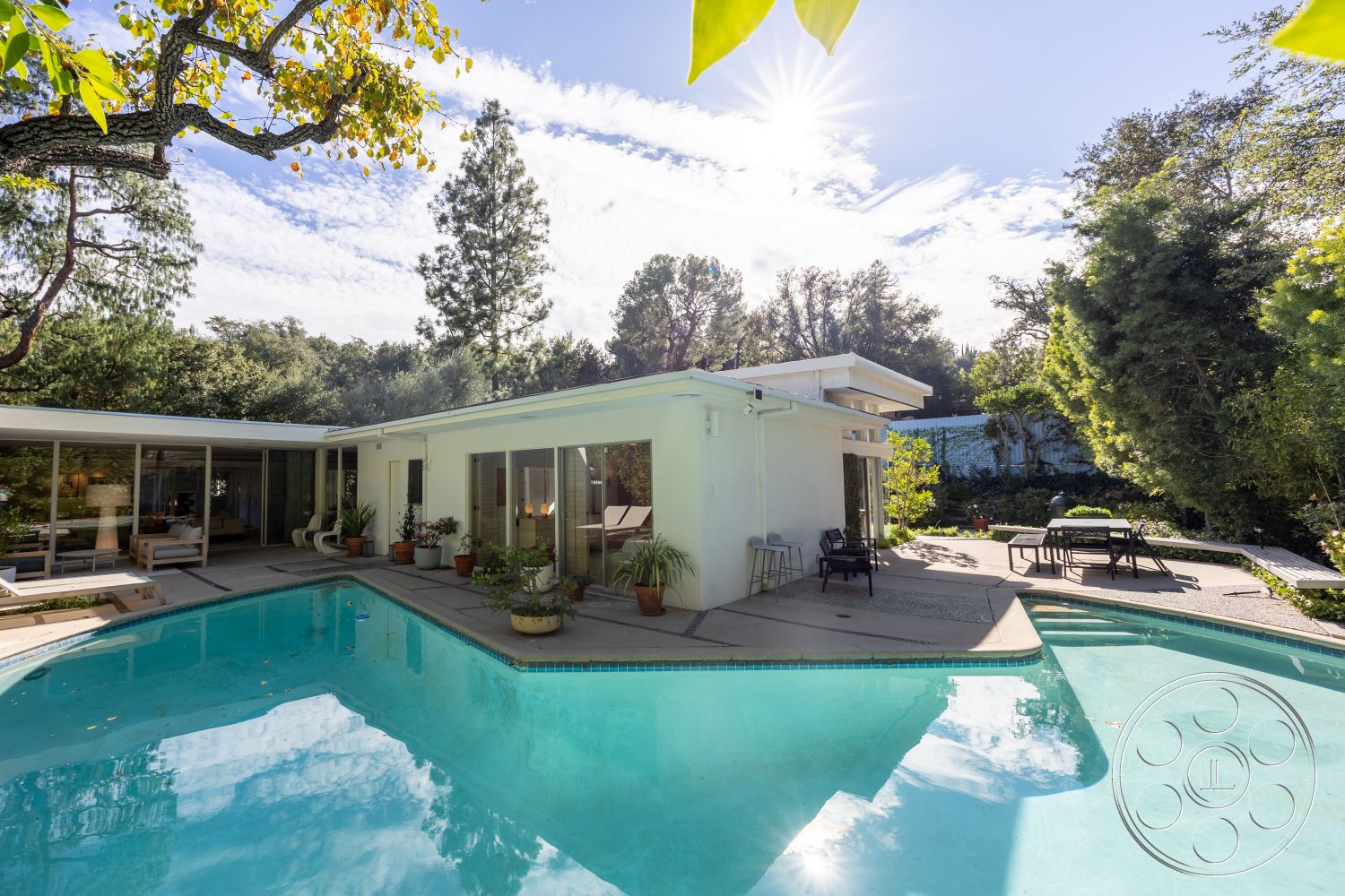 MCM 27 - rectangular swimming pool, outdoor dining area, landscaped backyard, modern architectural style, concrete pool deck, surrounded by trees, casual outdoor seating, concrete patio flooring, exterior white stucco walls, urban garden setting, floor-to-ceiling glass windows, sunlit outdoor space, clear blue sky view, sliding glass patio doors, potted plants decor, mid-century modern home, sunny exterior environment, hardwood outdoor lounge area, wooden garden furniture, poolside sun loungers