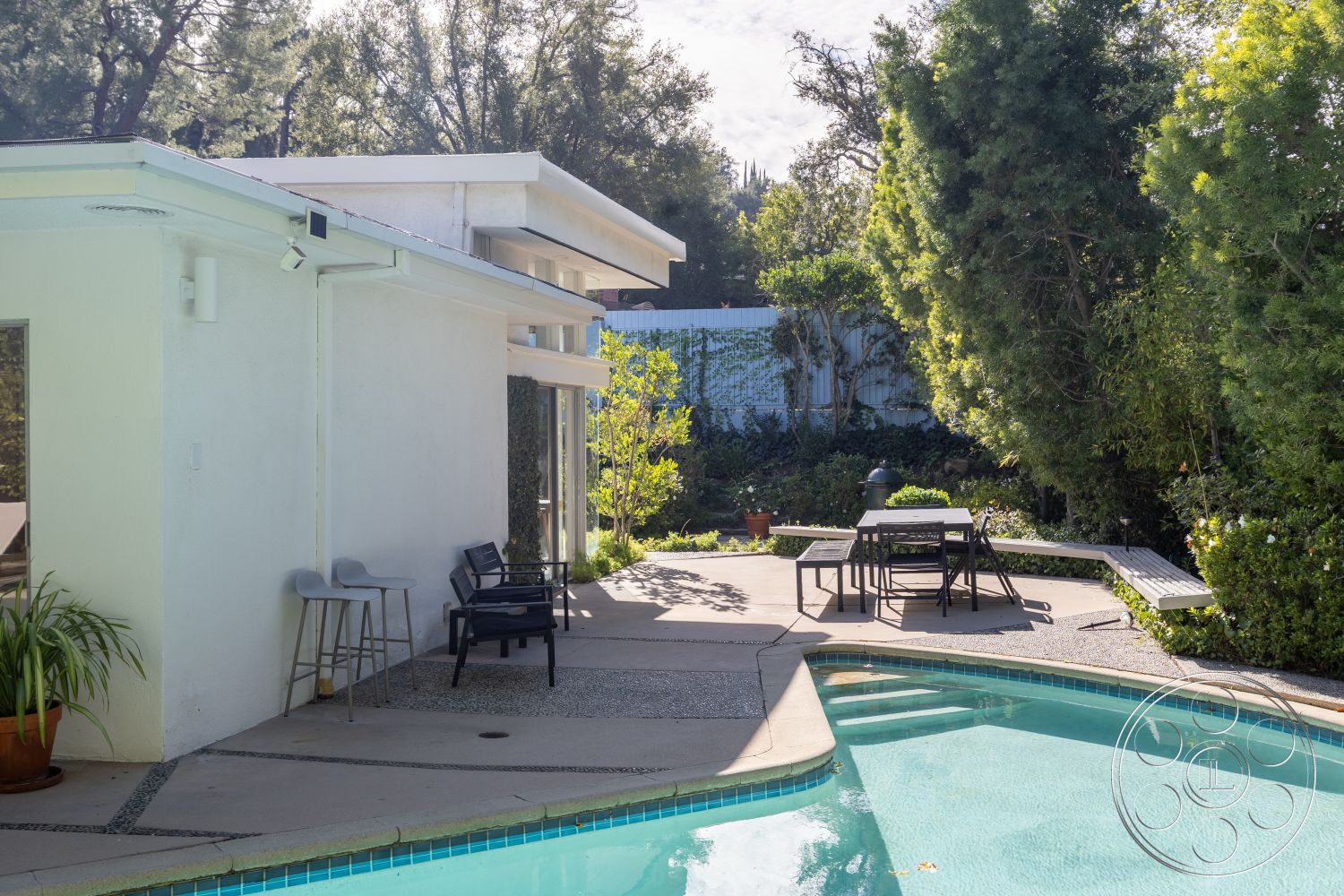 MCM 27 - exterior, white stucco walls, natural lighting, tropical plants, outdoor dining area, contemporary outdoor furniture, urban oasis, rectangular pool, glass patio doors, functional outdoor space, sunny backyard, landscaped garden setting, sliding glass door access, concrete steps leading to pool, concrete pool border, modern minimalist architecture, exposed aggregate concrete surface, steel outdoor railing, pebble finish patio flooring