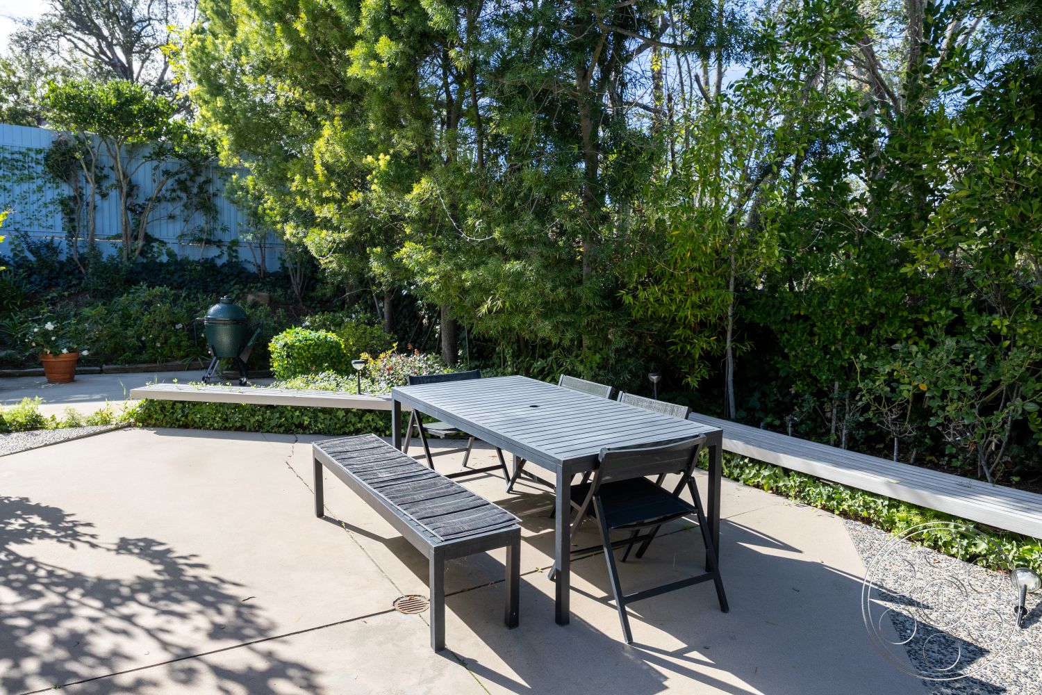 MCM 27 - gravel pathway, landscaped backyard, wooden bench seating, metal outdoor chairs, concrete patio flooring, natural light exposure, urban garden setting, exterior patio area, rectangular outdoor dining table, decorative flower pots, sloped garden bed, garden setting with lush greenery, modern outdoor dining space, surrounding plant life