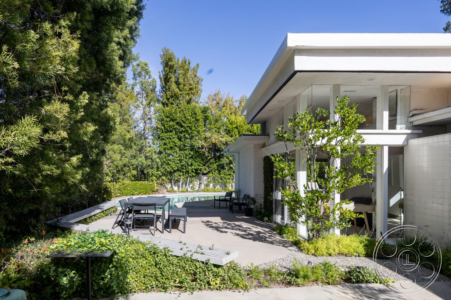 MCM 27 - concrete walkway, open-concept design, concrete steps leading to pool, floor-to-ceiling glass windows, minimalist architectural style, exposed concrete wall accents, exterior concrete patio flooring, rectangular pool area, modern steel outdoor furniture, lush landscaped garden setting, planter bed with greenery, wood deck around the pool, circular outdoor dining space