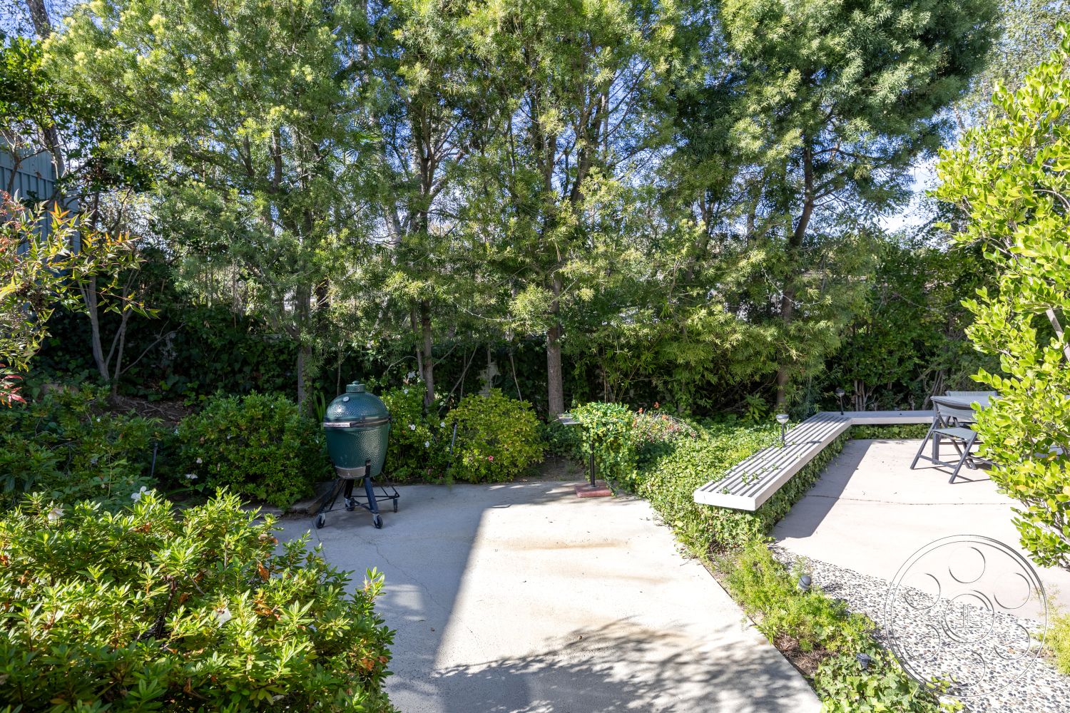 MCM 27 - wooden bench seating, natural stone pathway, contemporary outdoor design, lush greenery backdrop, landscaped garden setting, sunlit garden area, exterior patio area, concrete patio floor, green ceramic grill, gravel border landscaping, bushes and shrubs surrounding patio, rectangular outdoor dining space, seating area among trees