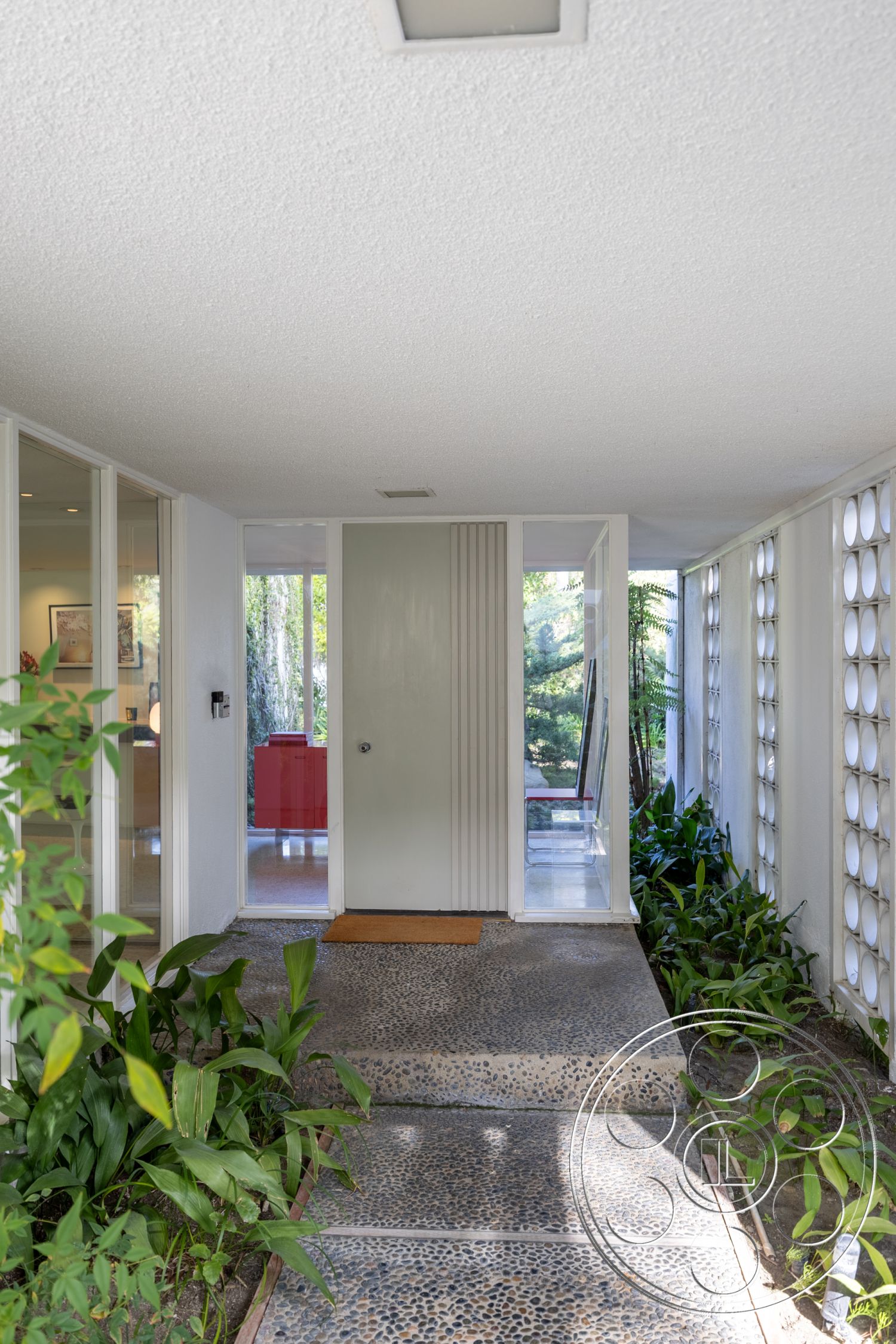 MCM 27 - clean lines, minimalist design, large windows for natural light, natural landscaping, textured plaster ceiling, organic shapes, contemporary aesthetic, open-concept layout, mid-century modern architecture, glass wall partitions, smooth concrete finish, modern front door, exposed aggregate concrete patio, pebble concrete walkway, lush green plants, interior entrance, greeting area