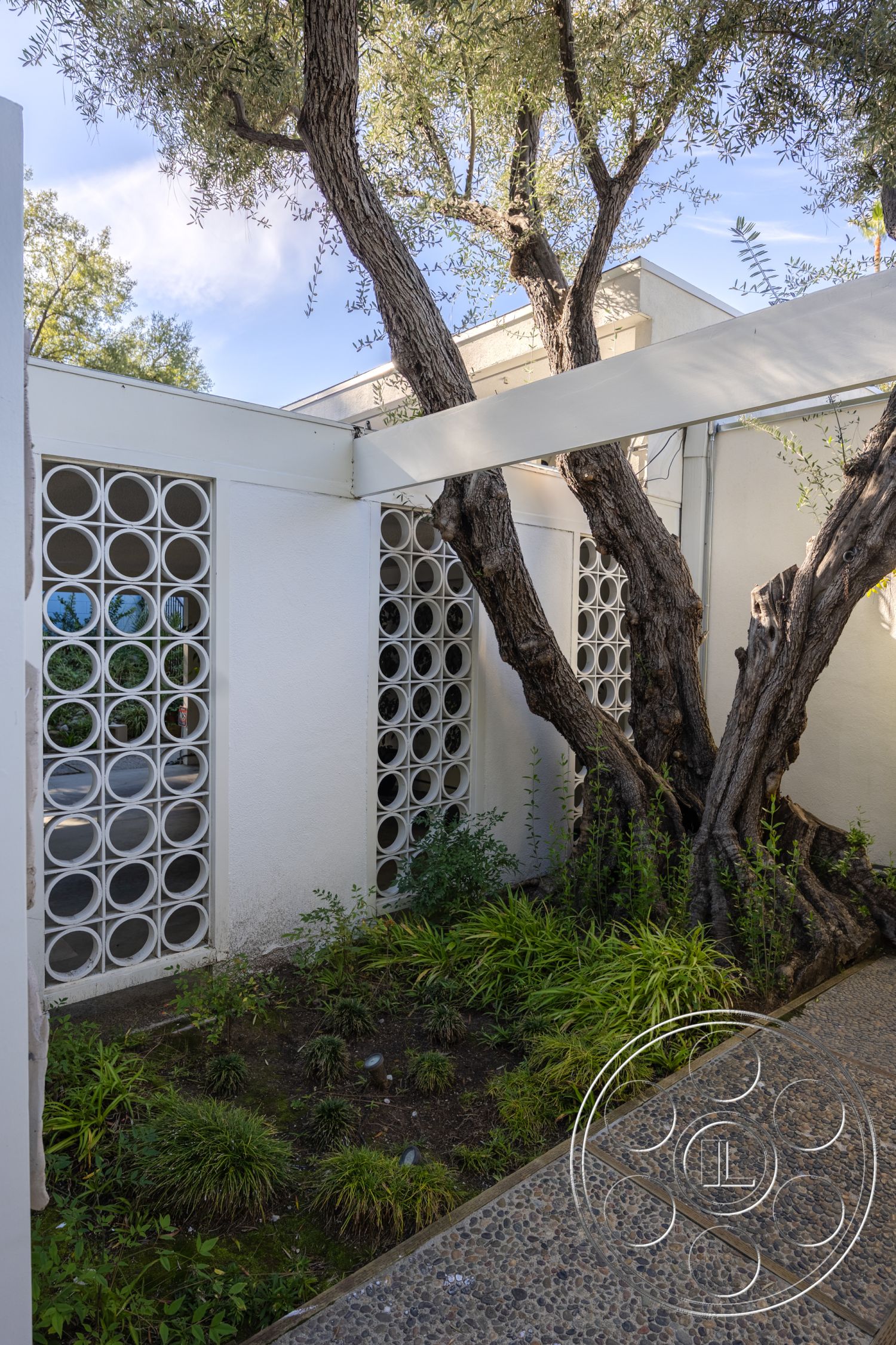 MCM 27 - exterior, modern architectural style, textured concrete wall, landscaped garden area, minimalist entryway design, native plantings, greenery surrounding structure, concrete garden pathway, decorative circular glass block wall, olive tree in garden, light natural stone gravel, horizontal wooden beams