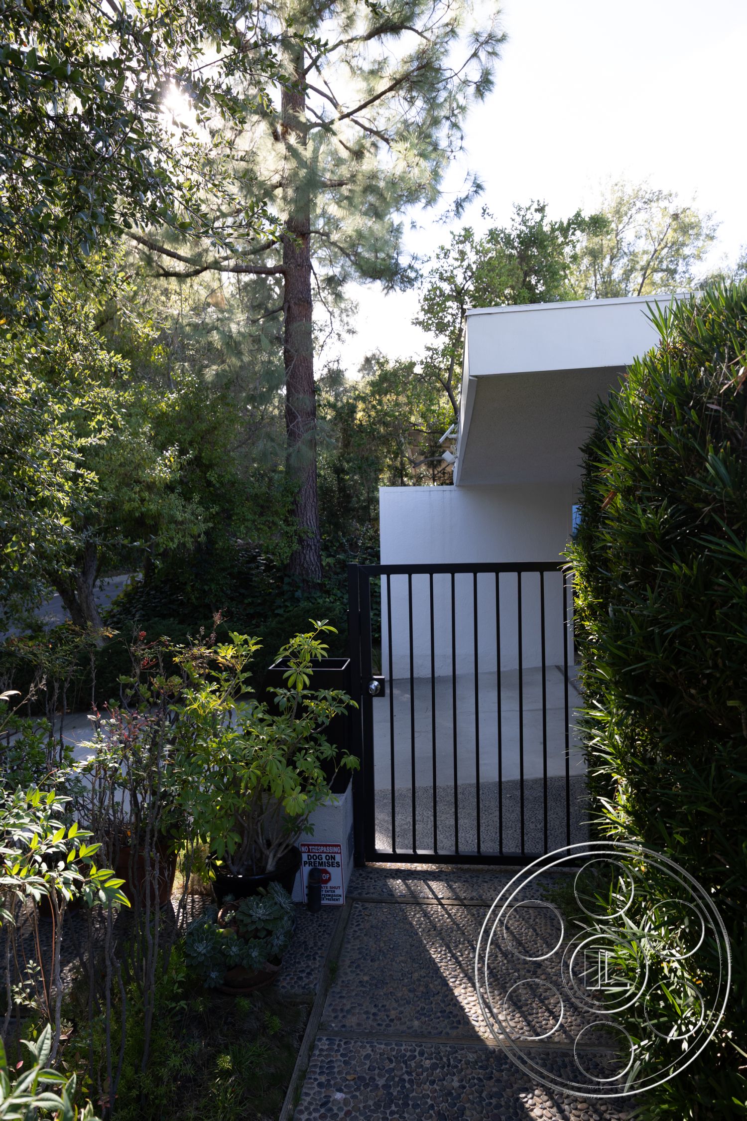 MCM 27 - exterior gate with black metal railing, pebble walkway with concrete stepping stones, dense greenery surrounding entrance, modern architectural design with flat roof, minimalist landscaping with low shrubs, direct access to driveway with smooth concrete surface, sunlit environment with dappled light through trees, pathway leading to garage area, tropical plant presence in landscape design, clear blue sky with bright daylight