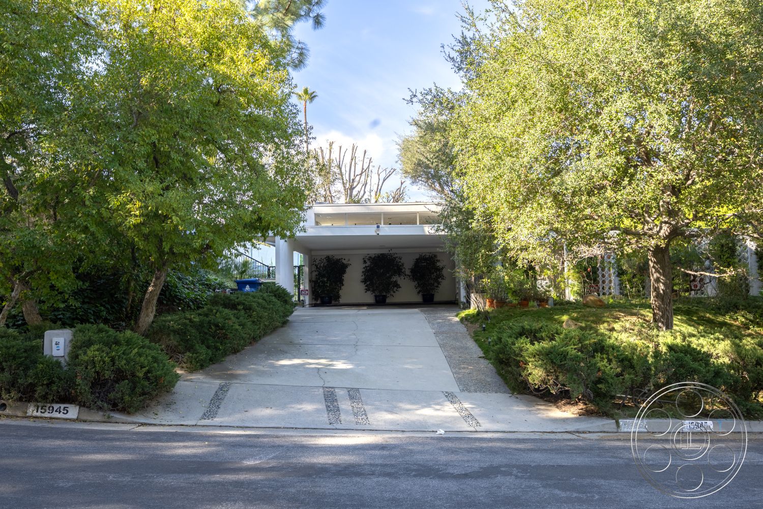 MCM 27 - modern architectural style, modern minimalist aesthetic, landscaped garden with native plants, lush greenery surrounding property, concrete carport structure, exterior concrete driveway with decorative stone inlay, flat roof design with overhangs, interior open-plan layout (implied), paved walkway flanked by hedges, tall ornamental trees providing shade, suburban setting with low-profile landscaping, invisible window design for natural light, low-maintenance exterior materials