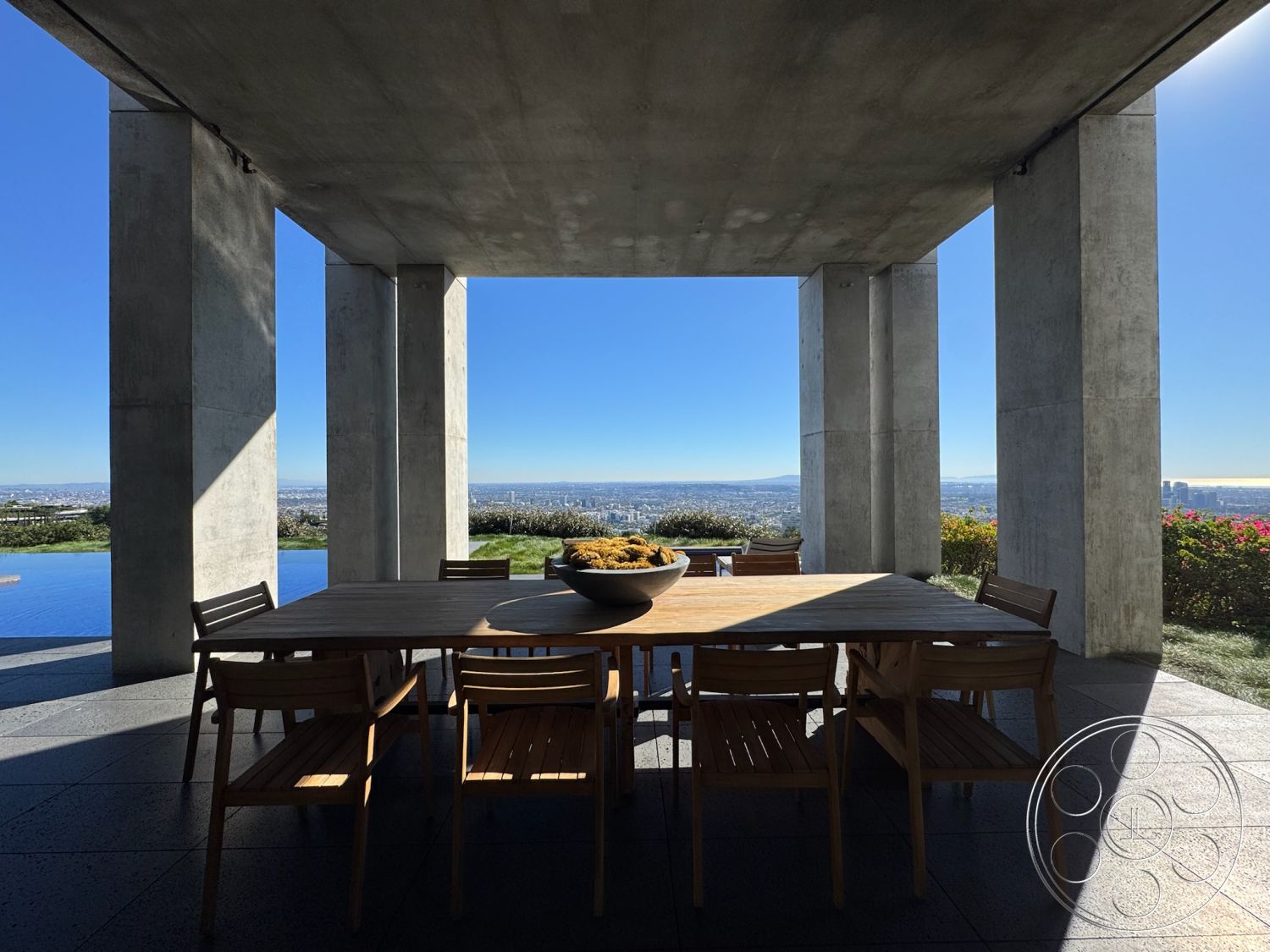 Fortress - minimalist design elements, concrete patio, modern architectural style, spacious outdoor area, panoramic city skyline view, wooden outdoor chairs, open-air dining space, wooden patio dining table, surrounding landscaped garden, exterior concrete support columns, concrete outdoor dining area flooring, infinity pool visible in background