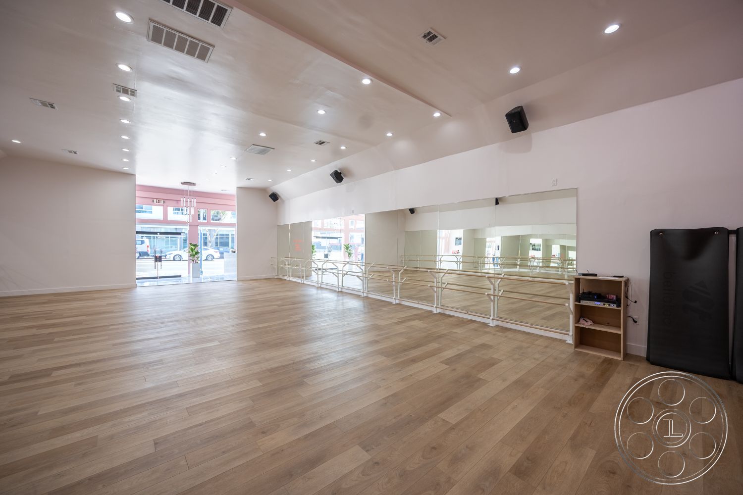 Gym 37 - open concept layout, urban setting, high vaulted ceiling, wooden storage cabinet, recessed lighting fixtures, acoustic ceiling panels, white painted interior walls, hardwood dance studio flooring, mirror wall installation, modern ballet bar railing, foyer entrance with large windows, commercial dance studio environment, natural light from storefront windows, black exercise mat, flexible studio space