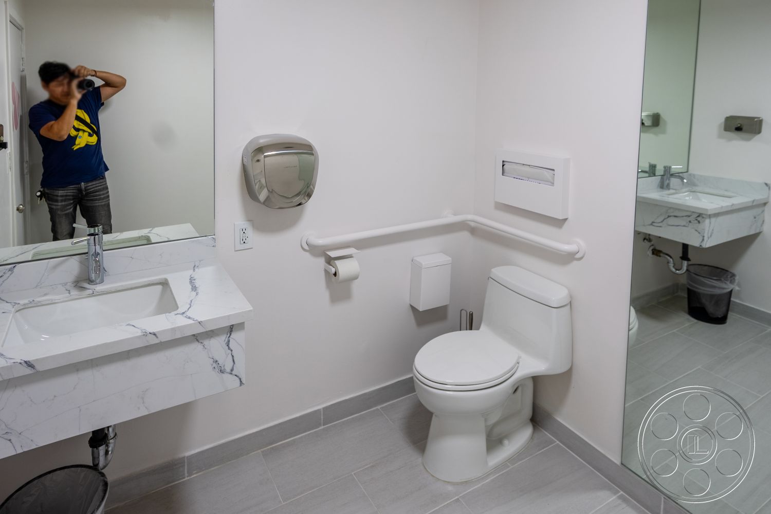 Gym 37 - rectangular bathroom mirror, exterior bathroom, marble countertop sink, wall-mounted soap dispenser, bright white painted walls, grab bars for accessibility, contemporary bathroom fixtures, porcelain toilet, textured gray tile flooring, flush-mounted toilet paper holder, minimalistic bathroom design, under-sink plumbing exposure, modern vanity with floating sink, wall-mounted paper towel dispenser