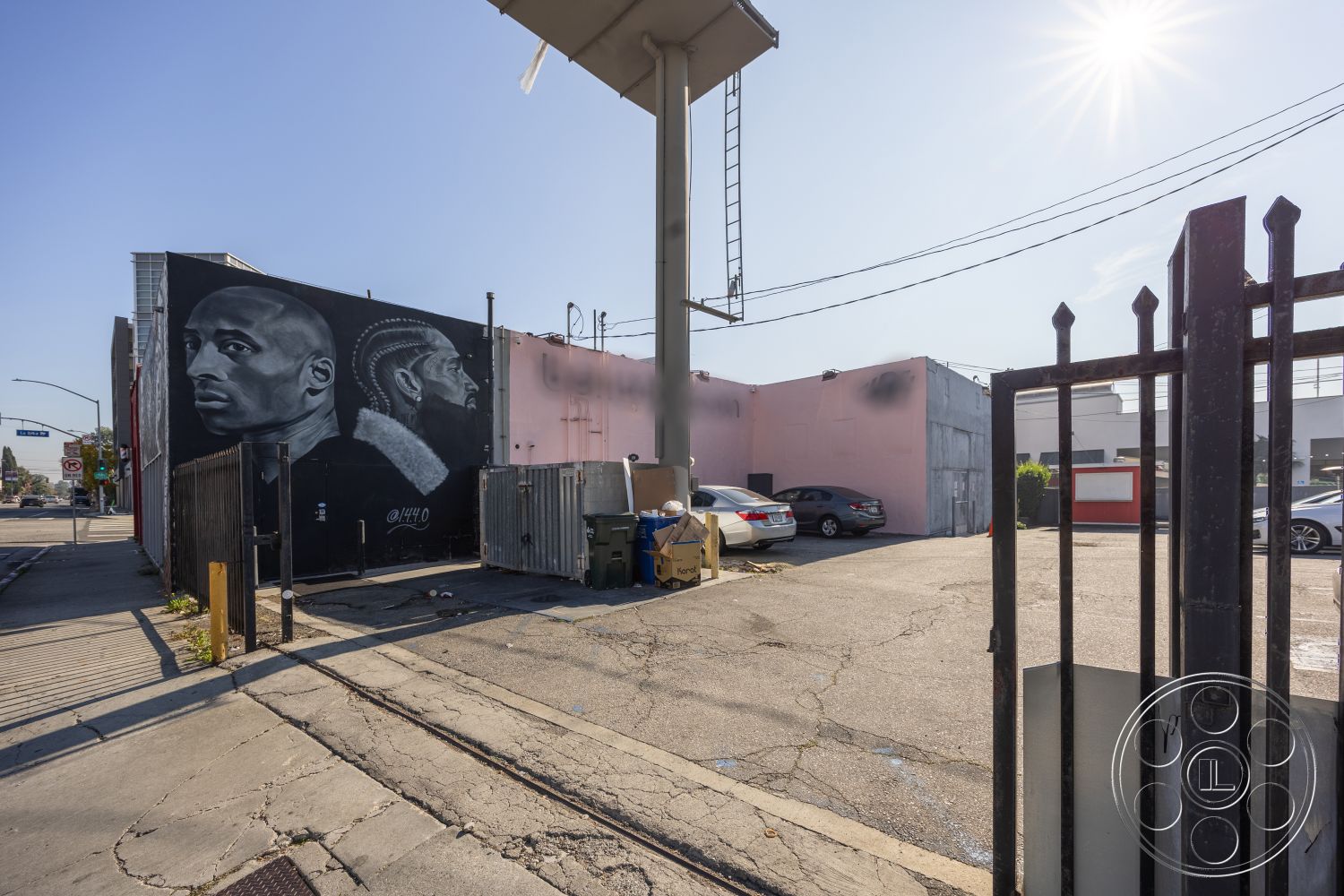 Gym 37 - exterior, urban setting, street view, urban environment, asphalt street, graffiti art wall, commercial property, concrete parking lot, flat roof structure, city sidewalk, steel fence gate, concrete wall with mural, industrial building facade, weathered paint finish, metal ladder, parking area with trash bins