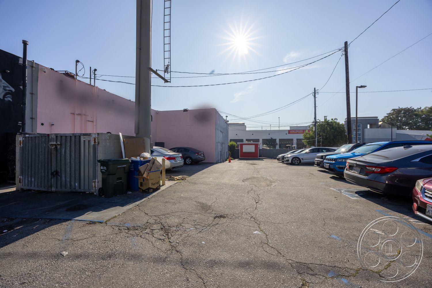 Gym 37 - exposed concrete walls, urban setting, functional outdoor space, asphalt parking lot, overhead power lines, industrial-style architecture, commercial building backdrop, metal dumpster, pink exterior wall, sunlight casting shadows, multiple parked vehicles, gravel debris on ground, flat pavement surface, curbside landscaping, utility pole details, colorful mural art