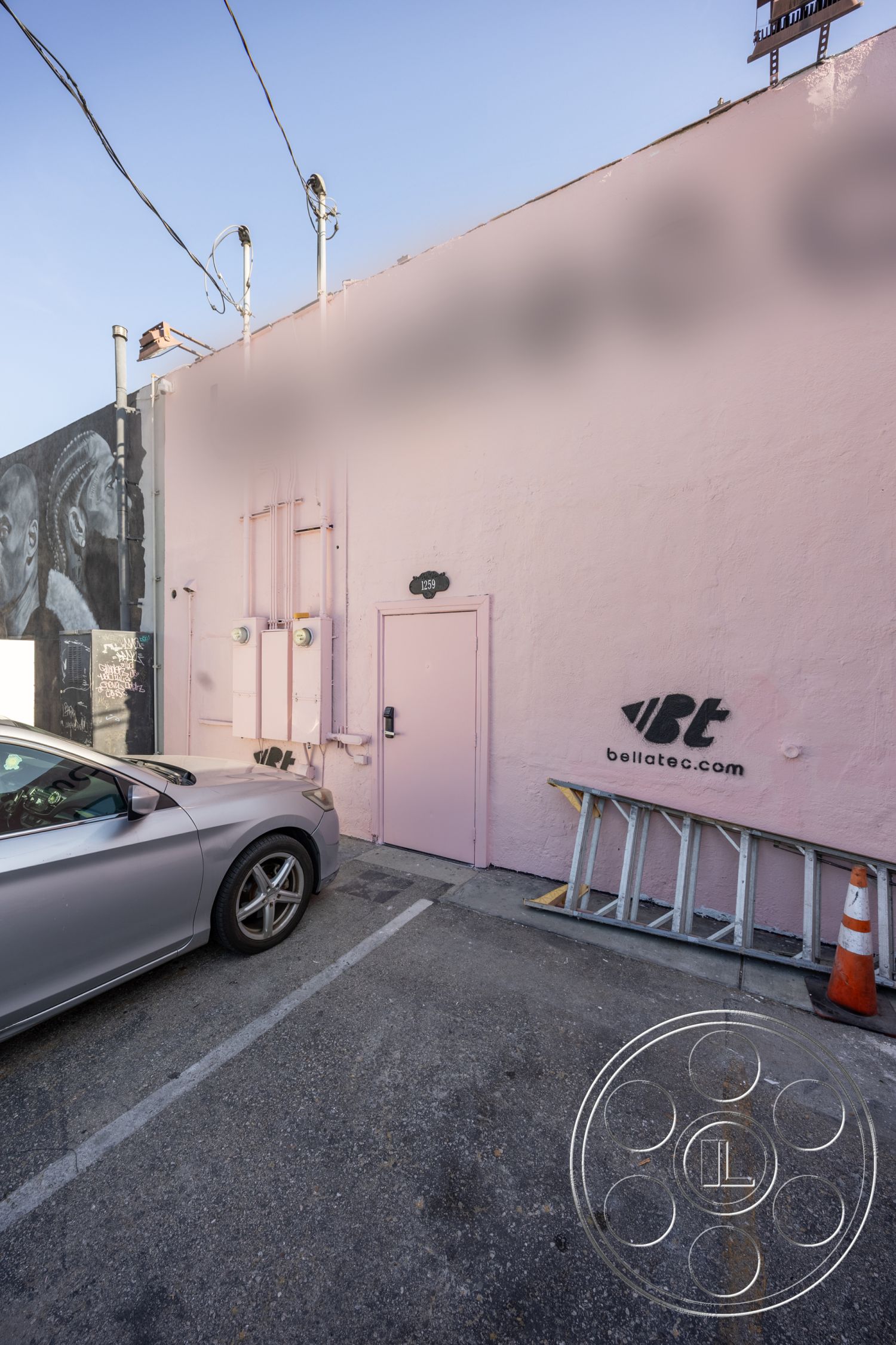 Gym 37 - orange traffic cone, concrete parking surface, modern commercial architecture, clear blue sky backdrop, exterior pink stucco wall, metal door with decorative handle, urban parking lot setting, graffiti art mural, industrial utility connections, signage detail on building, ladder against wall, single-story commercial space