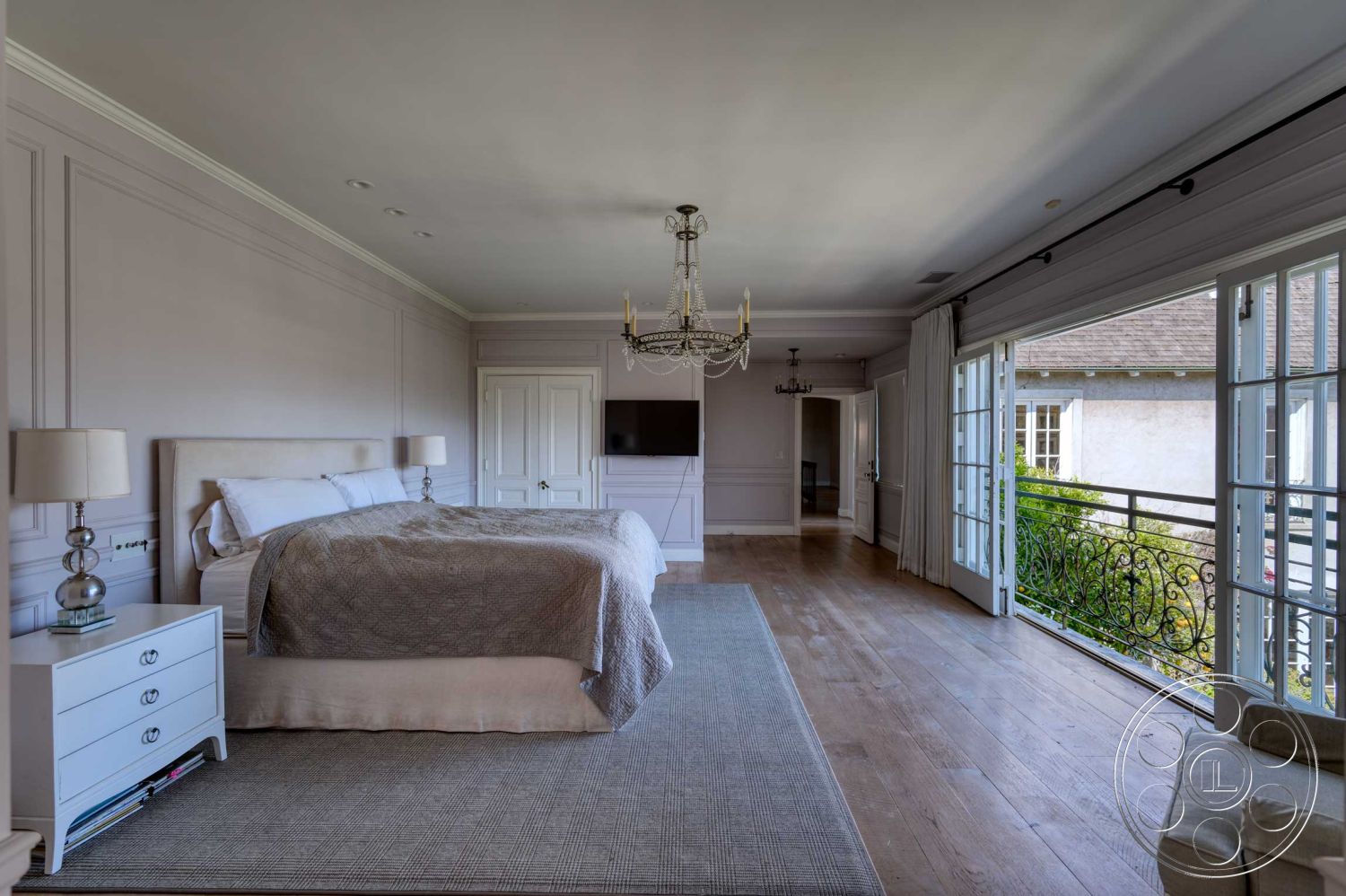 The French Chateau - textured plaster walls, hardwood bedroom floors, white painted walls, crown molding details, exterior balcony view, glass French doors, elegant bedside lamps, chandelier lighting fixtures, cohesive bedding composition, modern dresser with decorative elements