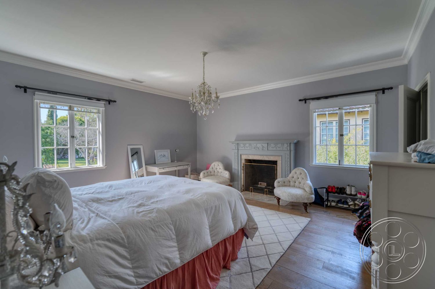 The French Chateau - textured area rug, decorative mirror, chandelier light fixture, exposed brick fireplace surround, hardwood bedroom floors, interior master bedroom, gray painted walls, white crown molding details, double hung windows, modern upholstered armchairs