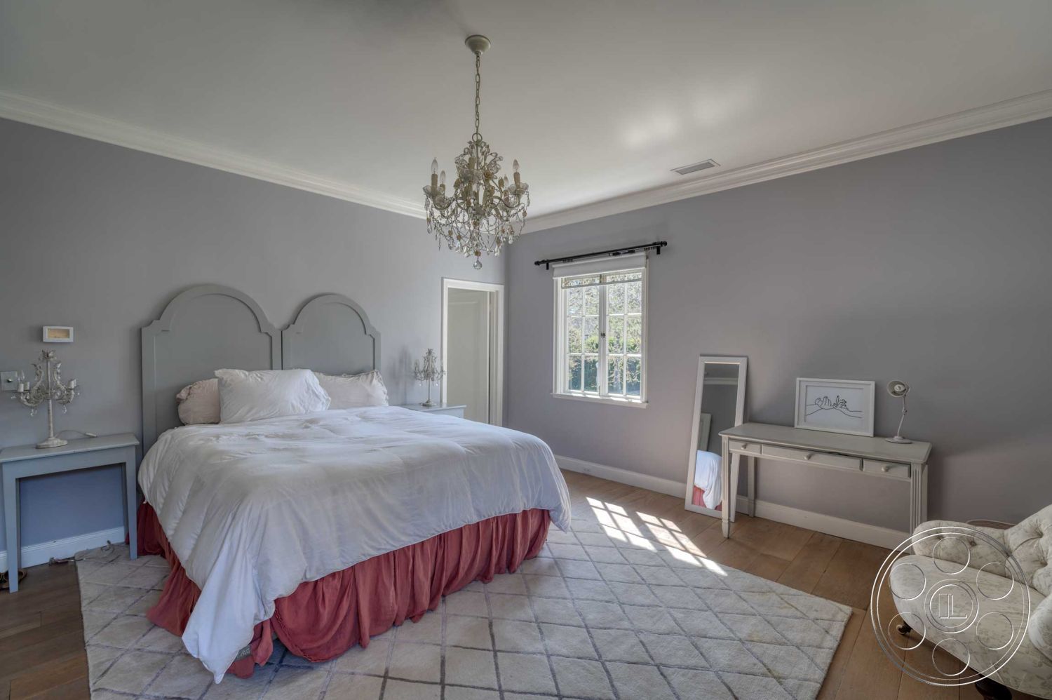 The French Chateau - textured area rug, wooden bedside table, elegant window treatments, interior bedroom, hardwood bedroom floors, minimalist decor style, gray painted walls, chandelier lighting fixture, exposed crown molding details, natural lighting from window, white upholstered bed, dressing table with mirror, plush upholstered armchair