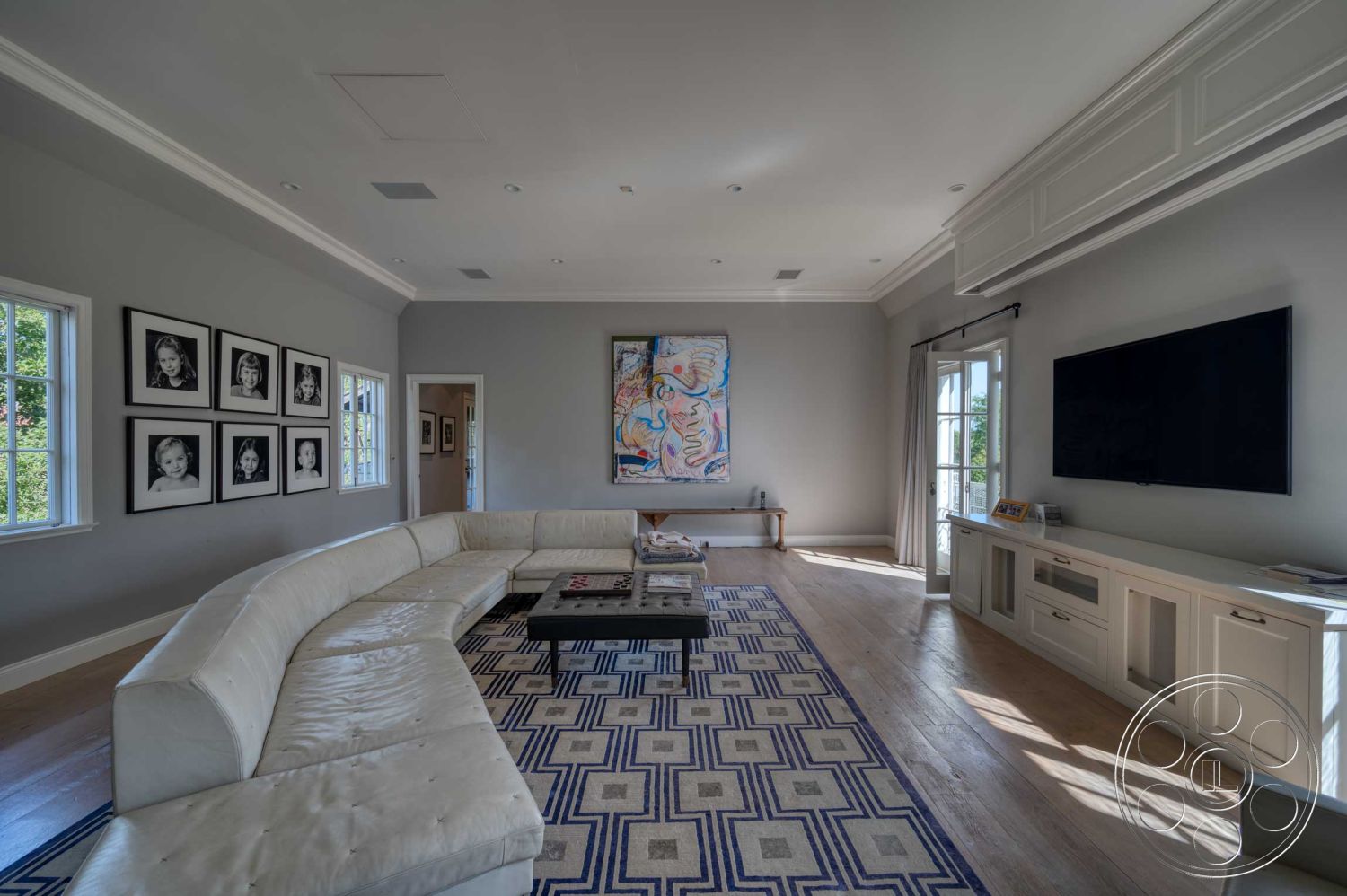 The French Chateau - neutral color palette, hardwood flooring, abstract wall art, textured walls, floor-to-ceiling windows, geometric area rug, black coffee table, curved sectional sofa, modern minimalist design, interior living room, soft beige curtains, white crown molding details, polar white painted walls, gallery wall with black and white framed photographs, built-in media console, natural light bright space