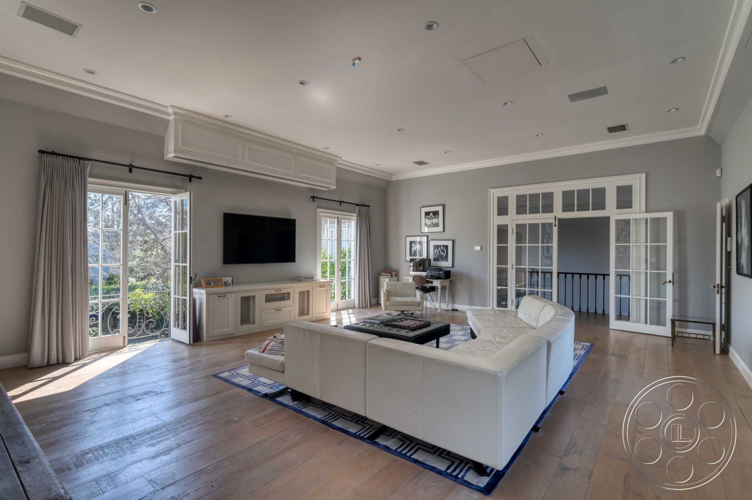 The French Chateau - neutral color palette, natural light from large windows, balcony view, glass patio doors, contemporary coffee table, white painted walls, modern sectional sofa, open floor plan interior, crown molding details, exposed ceiling details, light hardwood living room floors, black and white framed art on walls, contemporary entertainment unit