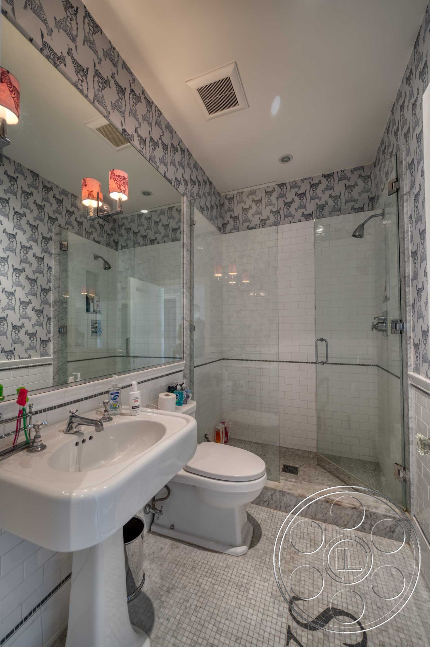 The French Chateau - recessed lighting, glass shower enclosure, pedestal sink, framed mirror, chrome faucet, contemporary light fixtures, glass shower doors, interior bathroom, marble bathroom flooring, modern toilet, white subway tile shower walls, marble mosaic flooring, decorative wallpaper with animal print, pink patterned lampshades, white baseboard trim, striped wall trim, silver towel rack