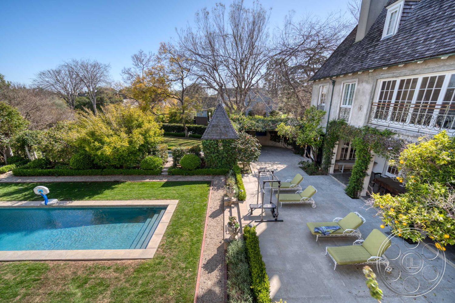 The French Chateau - rectangular swimming pool, stone pathway, outdoor living space, urban residence, poolside lounge area, hedge landscaping, landscaped garden area, exterior concrete pool border, exterior stucco walls, exterior concrete patio flooring, lush greenery surrounding pool, lounge chairs on patio, shingle roof on house, multi-pane French windows, sunlight-drenched courtyard, side garden with foliage, seasonal tree canopy, family backyard retreat