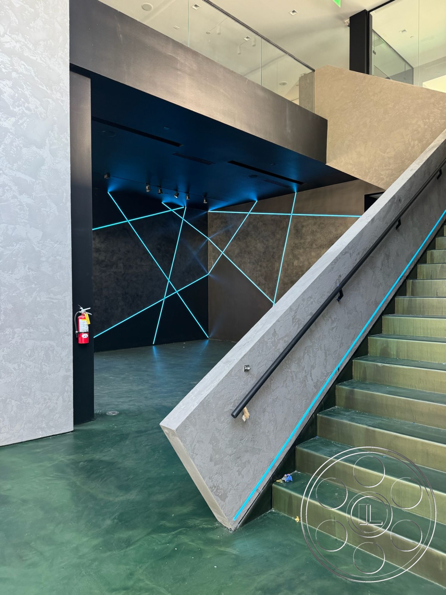 Sunset Studio - textured plaster wall treatment, interior polished concrete flooring, floor-to-ceiling windows providing natural light, exterior entrance with concrete steps, black accent wall with LED lighting, geometric pattern wall design with blue lighting, steel handrail on concrete staircase, open-concept space with high ceilings, modern architectural style with minimalist details, glass partition wall in entryway