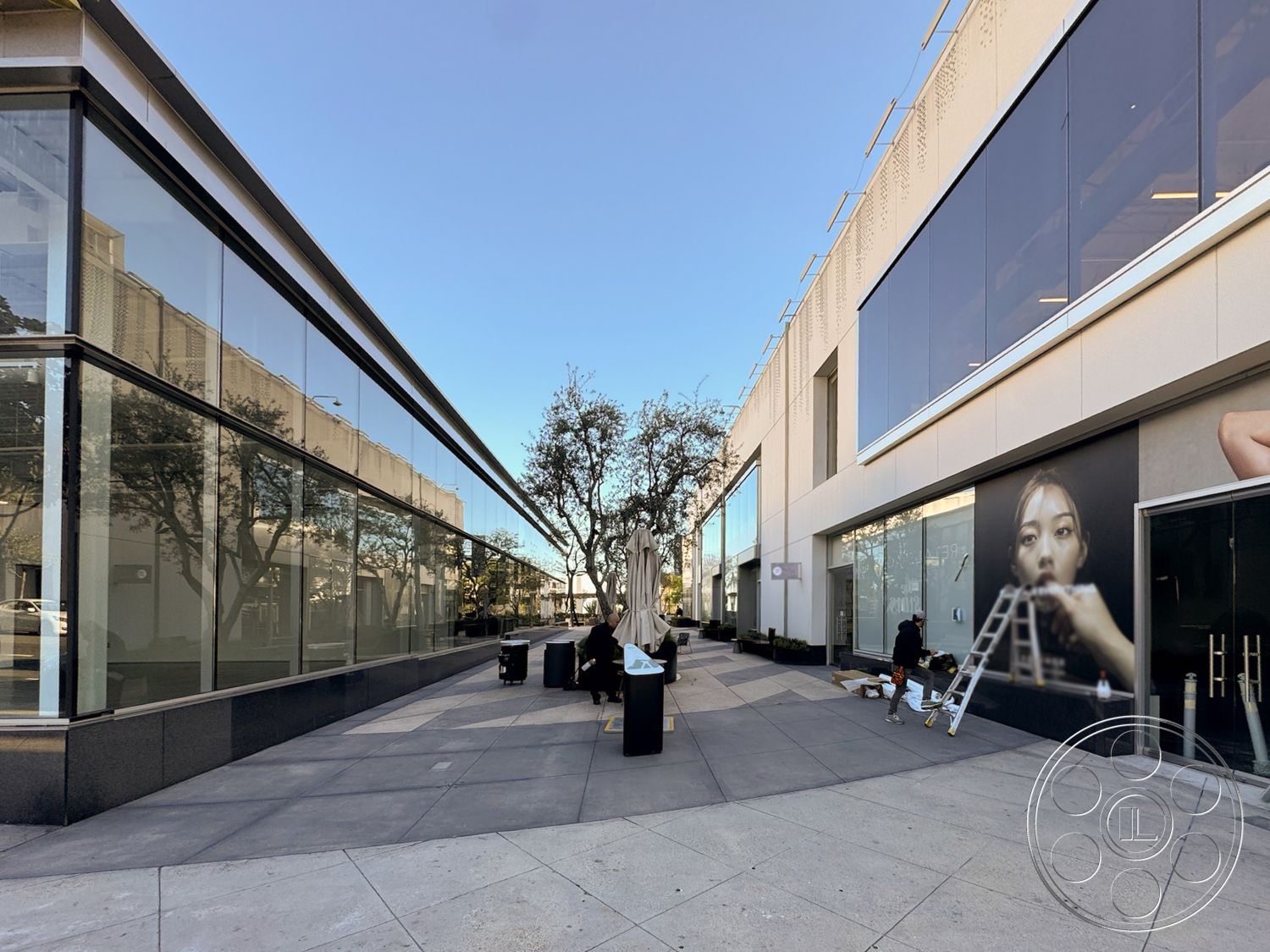 Sunset Storefront - outdoor seating area, modern architectural style, landscaped planters, large floor-to-ceiling windows, aluminum window frames, smooth plaster wall finish, bright sunny day setting, rectangular building layout, exterior glass storefronts, polished concrete sidewalk, granite base wall cap, tree-lined pedestrian walkway, urban shopping center environment, art mural wall feature, angled roof overhang, contemporary facade design, laminated glass panels, open-air gallery space, commercial retail space exterior