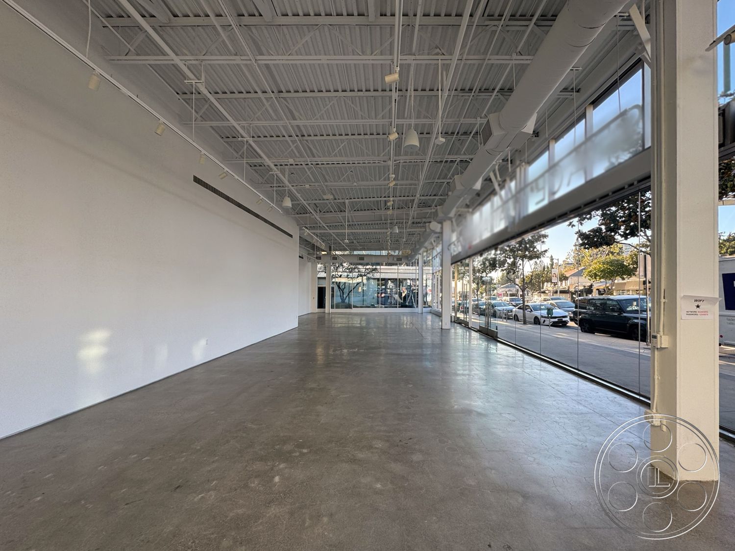 Sunset Storefront - commercial space, open plan layout, white painted walls, natural light exposure, interior concrete flooring, minimalist architectural style, large glass storefront windows, exposed steel structural beams, fluorescent ceiling lighting, high ceilings with metal grid framework, concrete finish walls, polished concrete floor finish, urban storefront setting, flexible space design