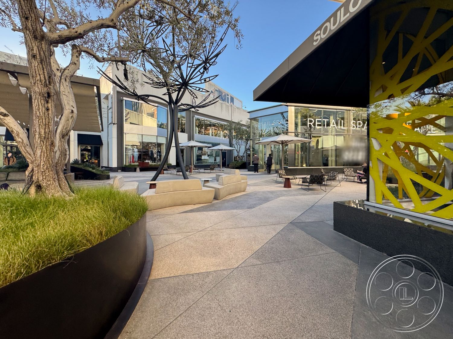 Sunset Storefront - exterior, contemporary design aesthetic, large storefront windows, surrounding urban environment, polished concrete plaza flooring, curved bench seating, modern angular architectural forms, metal tree sculpture installation, outdoor seating area with umbrellas, green grass landscaping in planters, black metal planter boxes, glass storefront facade, abstract geometric design elements, urban retail space setting, sunlit outdoor gathering space