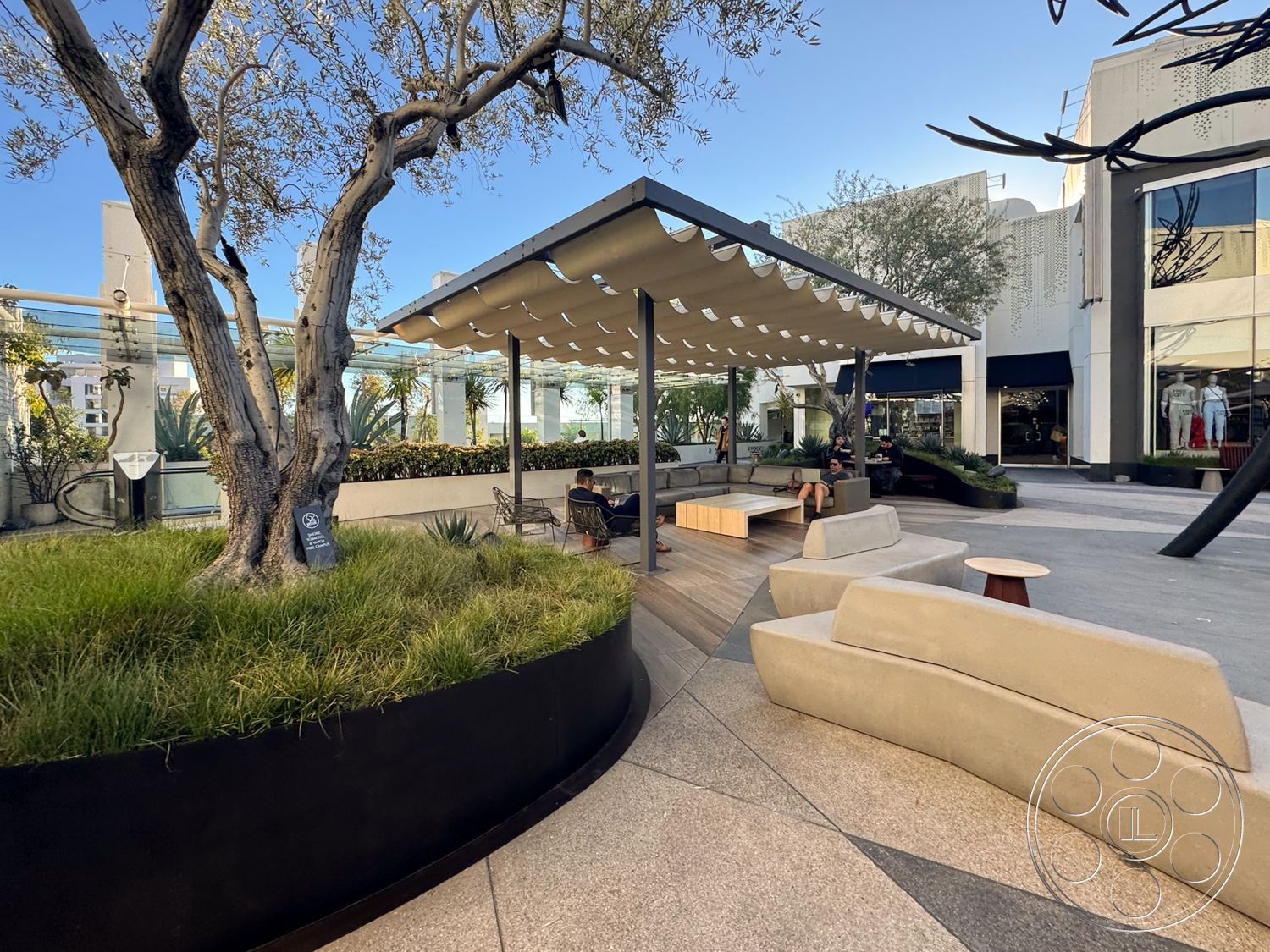 Sunset Storefront - exterior space, city skyline view, concrete patio flooring, minimalist outdoor furniture, modern lounge seating, landscaped planter beds, pergola with fabric canopy, curved metal flower beds, natural grass ground cover, wood deck area, decorative tree planting, urban outdoor seating, softscape landscaping features