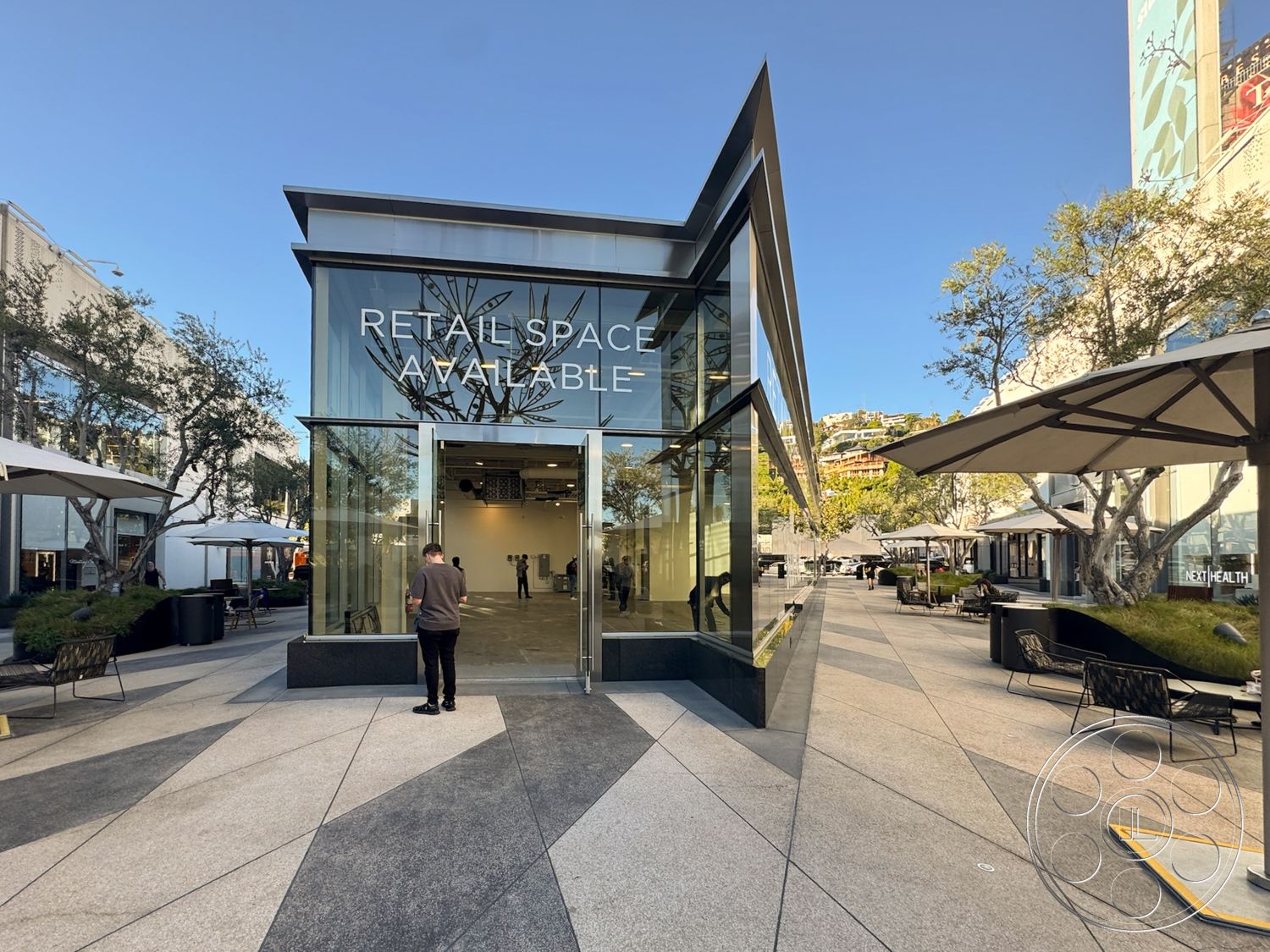 Sunset Storefront - modern architectural style, outdoor patio space, flat roof design, open floor plan interior, floor-to-ceiling glass walls, steel frame construction, concrete paver walkway, urban plaza setting, natural sunlight exposure, landscaped outdoor seating area, transparent glass doors, exterior retail space, glass storefront, polished granite base, triangular roof shape, umbrella-covered seating, geometric facade details