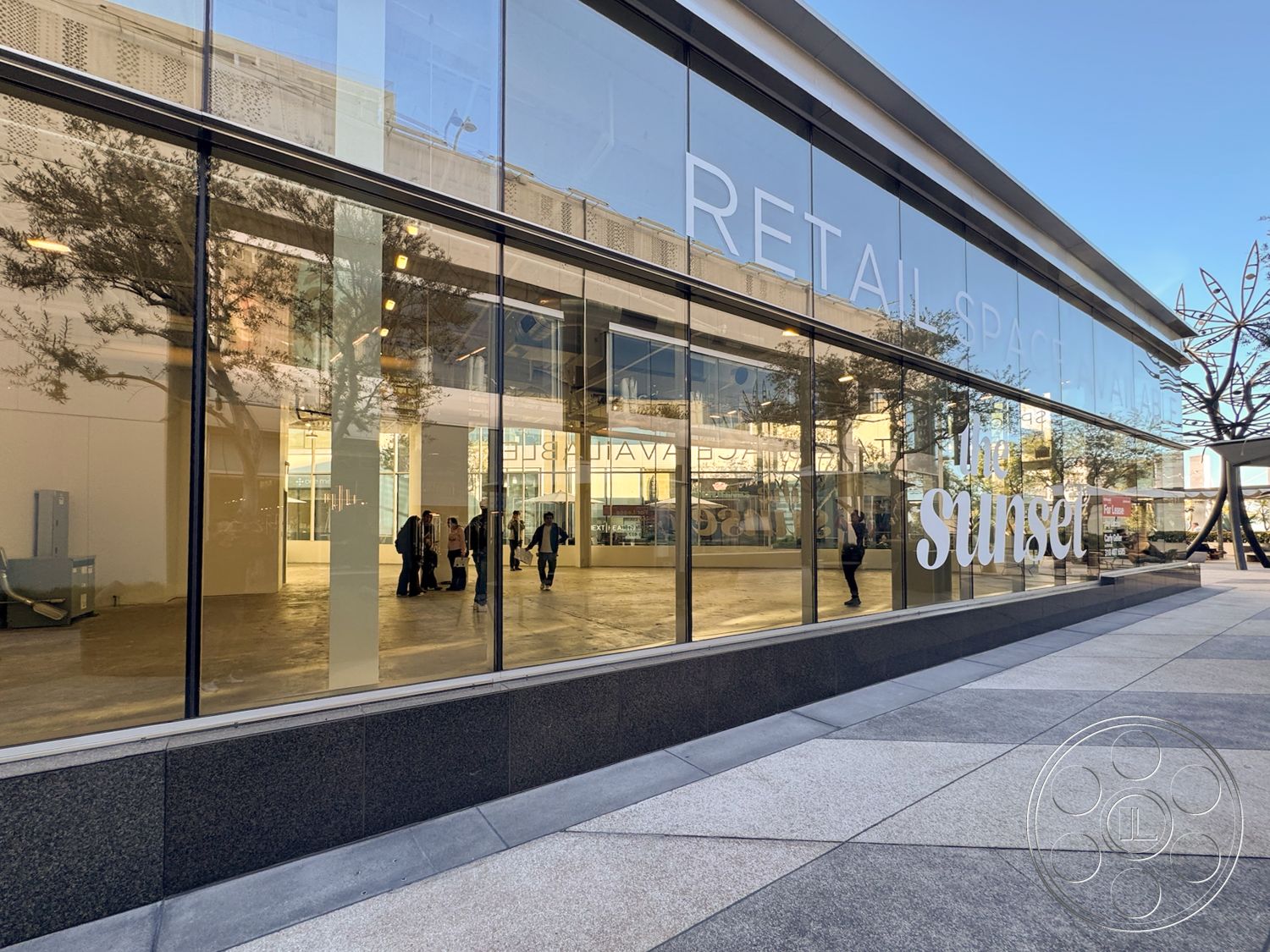 Sunset Storefront - polished concrete floor, steel framework, integrated landscaping, city street view, large glass panels, concrete sidewalk, open commercial space, modern architectural design, rectangular building shape, textured stone base, daylight illumination, exterior glass storefront, urban retail space, minimalistic façade, large entrance glass doors, art installation in plaza