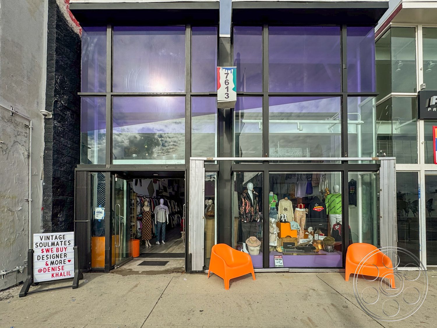 Melrose Store - minimalist seating, large floor-to-ceiling windows, concrete sidewalk, modern architectural details, exterior storefront design, urban retail environment, urban commercial setting, glass entrance doors, black textured wall finish, purple tinted glass paneling, angled roof structure, bright orange plastic chairs, vintage display signage, open entrance layout, inviting retail exterior, colorful fashion displays