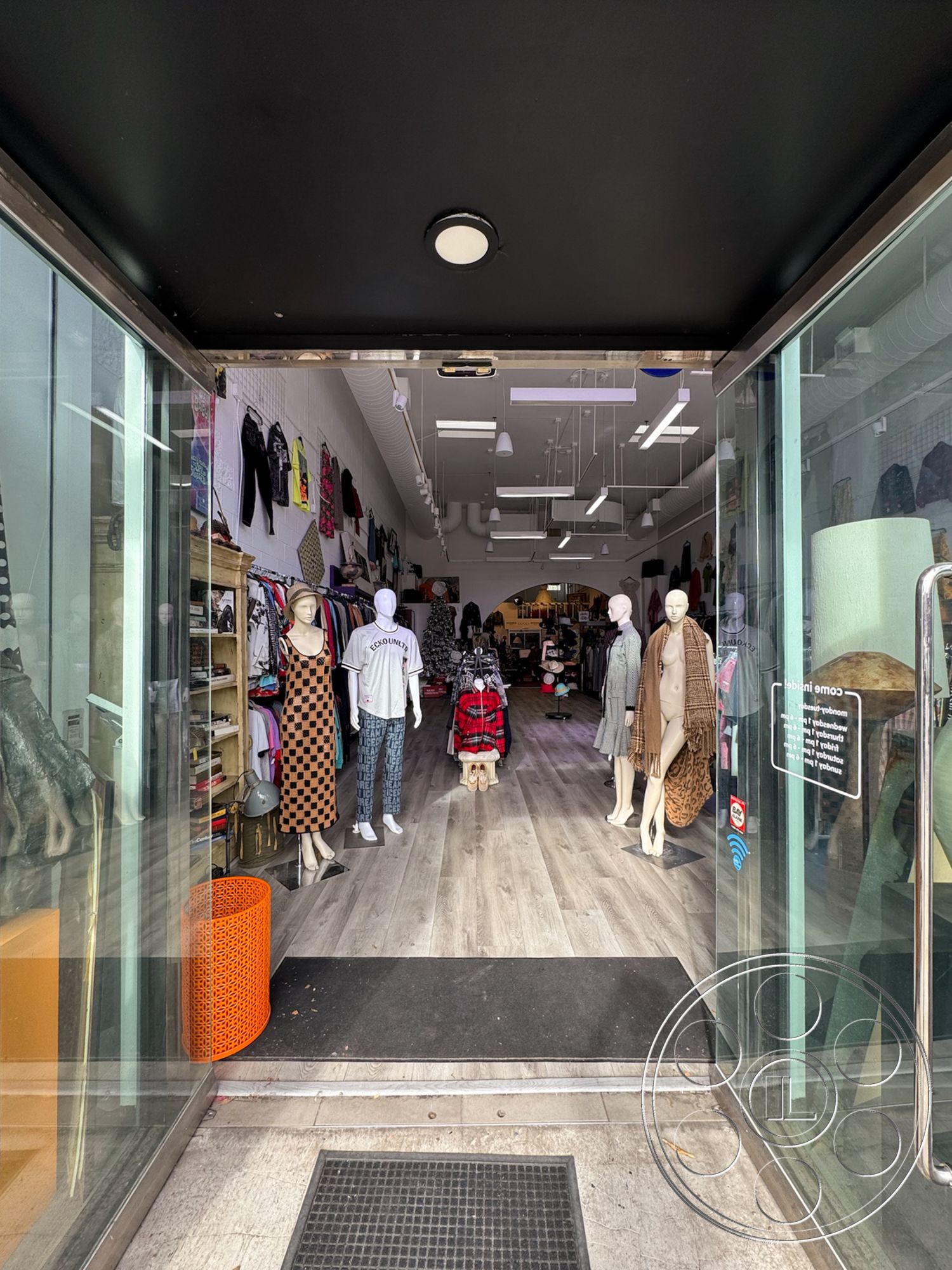 Melrose Store - minimalist aesthetic, textured wall finishes, metal light fixtures, open floor plan interior, wood laminate flooring, exposed ductwork ceiling, glass storefront entrance, black painted ceiling, colorful accent decor, modern retail layout, merchandise display racks, retail display mannequins, brightly colored accessories display, contemporary fashion boutique, sitting area with decorative stool, floor mats at entrance, clear glass entryway, storefront view of clothing line, dim lighting for ambiance, urban shopping district setting