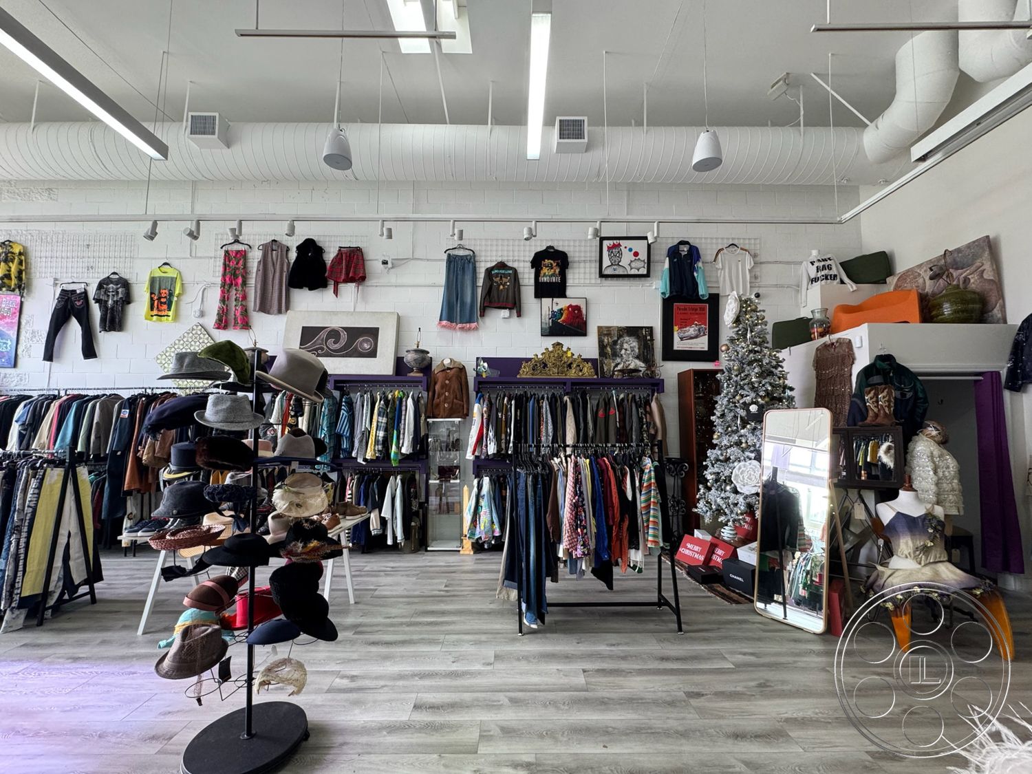 Melrose Store - open floor plan, decorative Christmas tree, plush seating area, interior retail space, vinyl wood plank flooring, metal clothing racks, urban boutique setting, exposed white brick walls, suspended ceiling with HVAC ducts, high ceiling with LED lighting, wall-mounted artwork displays, large mirror for customer use, colorful clothing displays, adjustable spotlights for merchandise, vibrant and eclectic decor, hats display stand, mannequin showcasing outfits, clothing labels and tags, fitting area with curtains