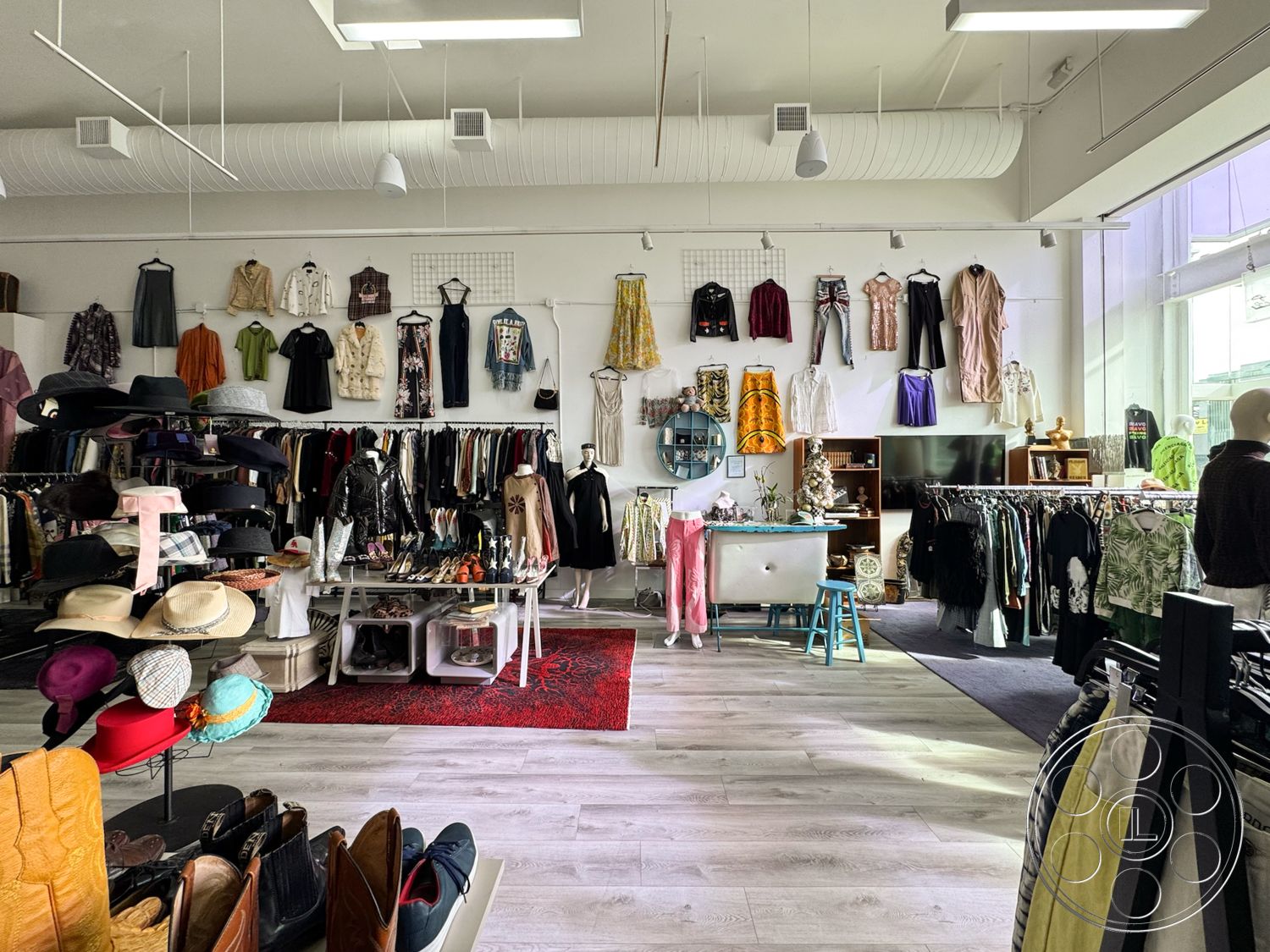 Melrose Store - decorative area rug, modern lighting fixtures, laminate flooring, stylish seating area, natural light through large windows, white painted walls, high ceilings with exposed ductwork, interior clothing boutique, display racks for clothing, vintage wooden shelving units, stacked hat display, mannequins displaying outfits, colorful clothing assortment, textured fabric hanging garments, retail merchandise displays, casual shopping environment