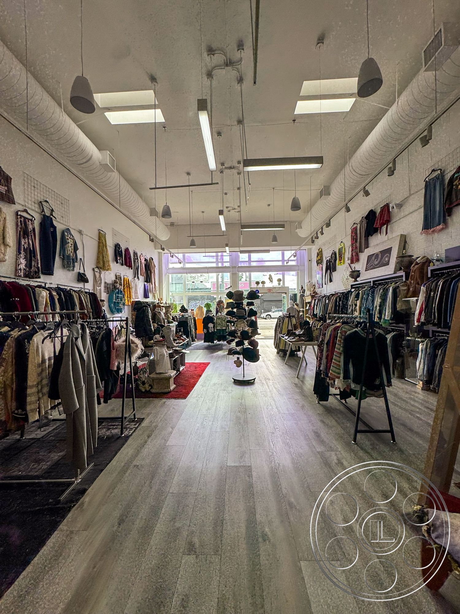 Melrose Store - natural light from skylights, exposed ductwork ceiling, textured white wall finish, interior clothing store, transformable storefront layout, light-colored wood plank flooring, open retail space design, multi-layered clothing display racks, modern industrial fixtures, vibrant colored clothing assortment, decorative display mannequin, modern shopping atmosphere, clothing rail displays, contemporary retail environment, clear glass storefront windows, large entrance area, focal point cash register area