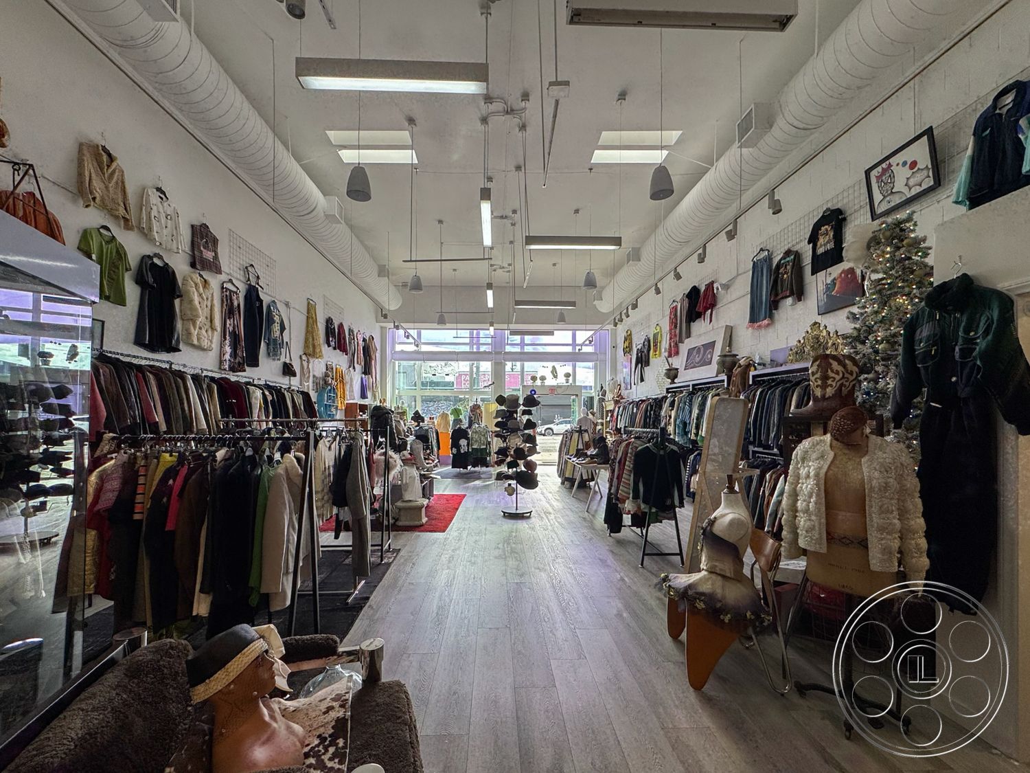 Melrose Store - hardwood flooring, open layout, artwork on walls, modern aesthetic, mirrored walls, white painted walls, large storefront windows, urban commercial setting, exposed ductwork ceiling, bright lighting fixtures, interior retail space, clothing display racks, mannequin displays, vintage clothing, decorative holiday tree, multiple fitting rooms, clothing boutiques, colorful garment displays, sitting area with plush furnishings