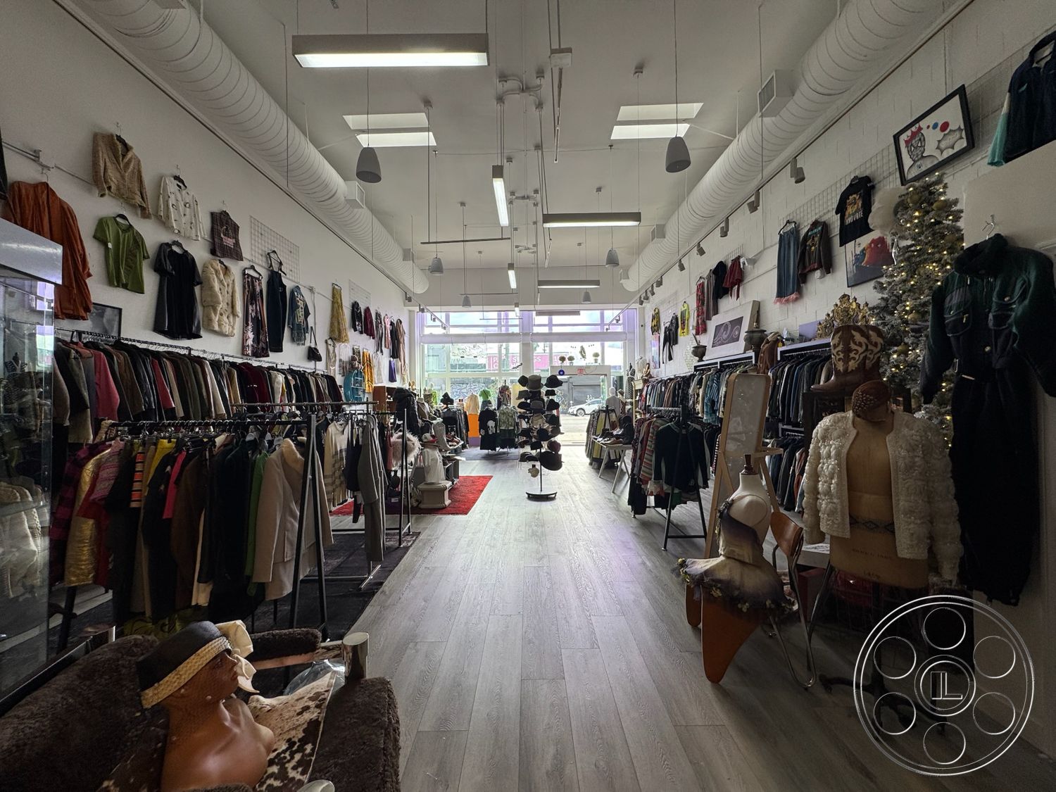 Melrose Store - decorative Christmas tree, laminate wood flooring, minimalist interior design, open floor plan layout, bright and airy ambiance, exposed ductwork ceiling, modern track lighting, interior retail space, white painted concrete walls, fitting room area, mannequins displaying clothing, multicolored clothing racks, window display with street view, cozy seating area with decorative pillows, textured wall art displays, stylish fashion items from various eras