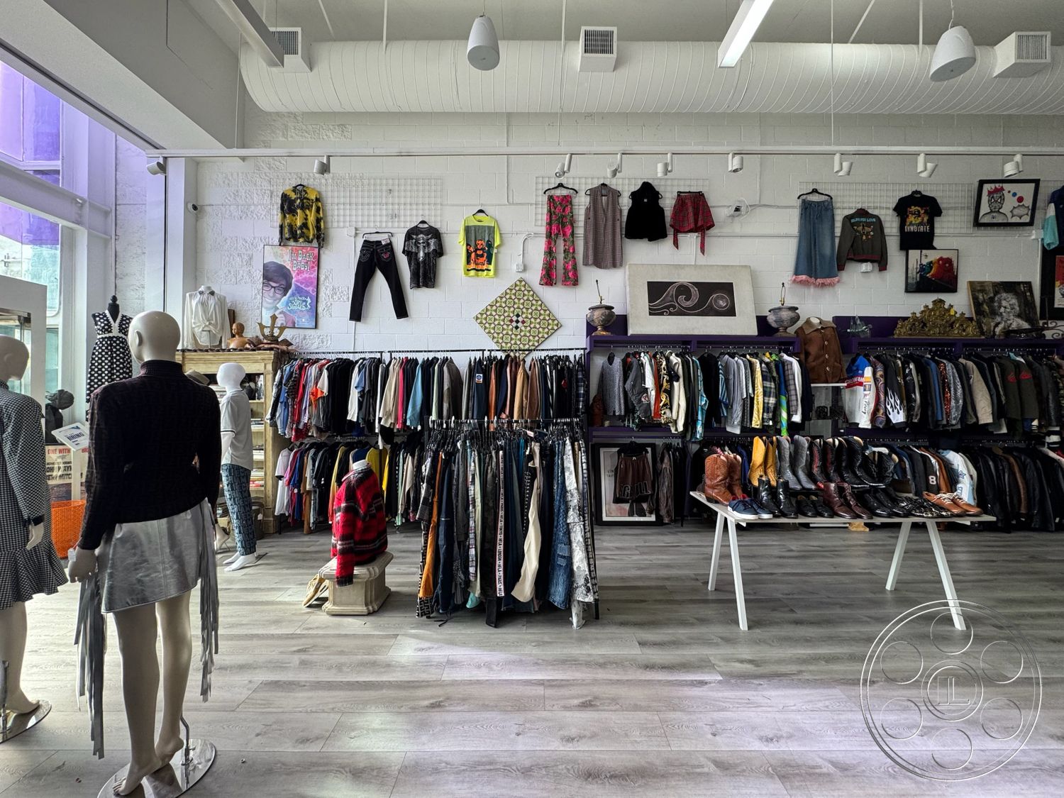 Melrose Store - open floor plan, artistic wall decor, laminate wood flooring, white painted walls, exposed brick accent wall, metal clothing racks, contemporary retail design, colorful clothing displays, casual shopping environment, interior shop space, mannequins displaying apparel, high ceiling with exposed ductwork, bright natural light from large windows, fashion merchandise arrangement, multifunctional retail space, urban boutique atmosphere, minimalist interior styling