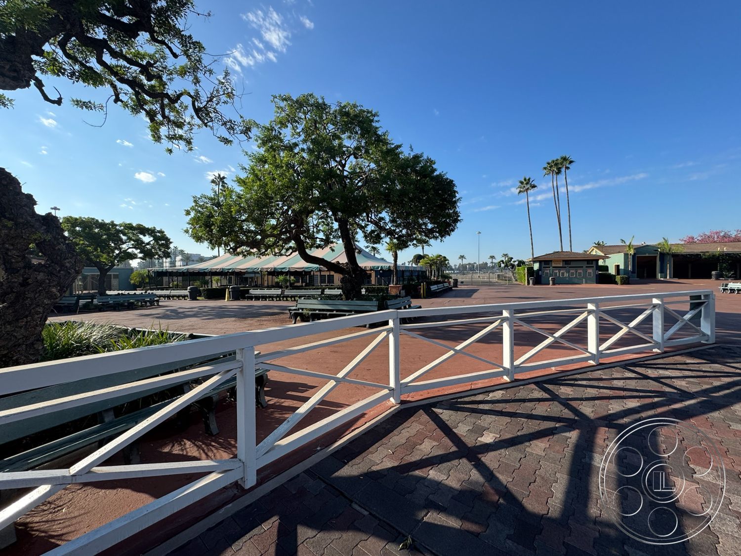 Arcadia Race Track - surrounding palm trees, casual outdoor gathering space, landscaped garden areas, exterior plaza setting, textured paver flooring, white wooden fence with X-bracing, large trees providing shade, outdoor seating area with tables, sunny blue sky background